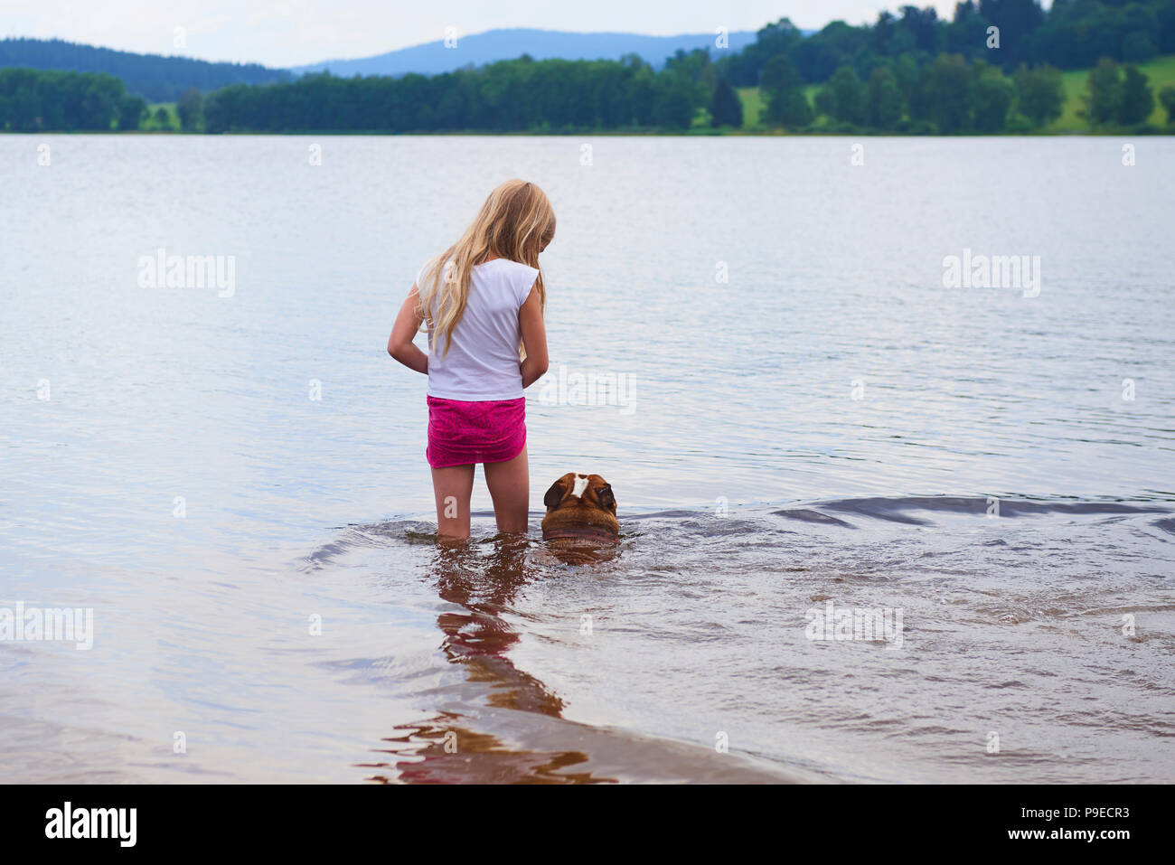 Kind blondes Mädchen in Shirt und Rock stehen im Wasser angezogen und das Spiel mit dem Hund Stockfoto