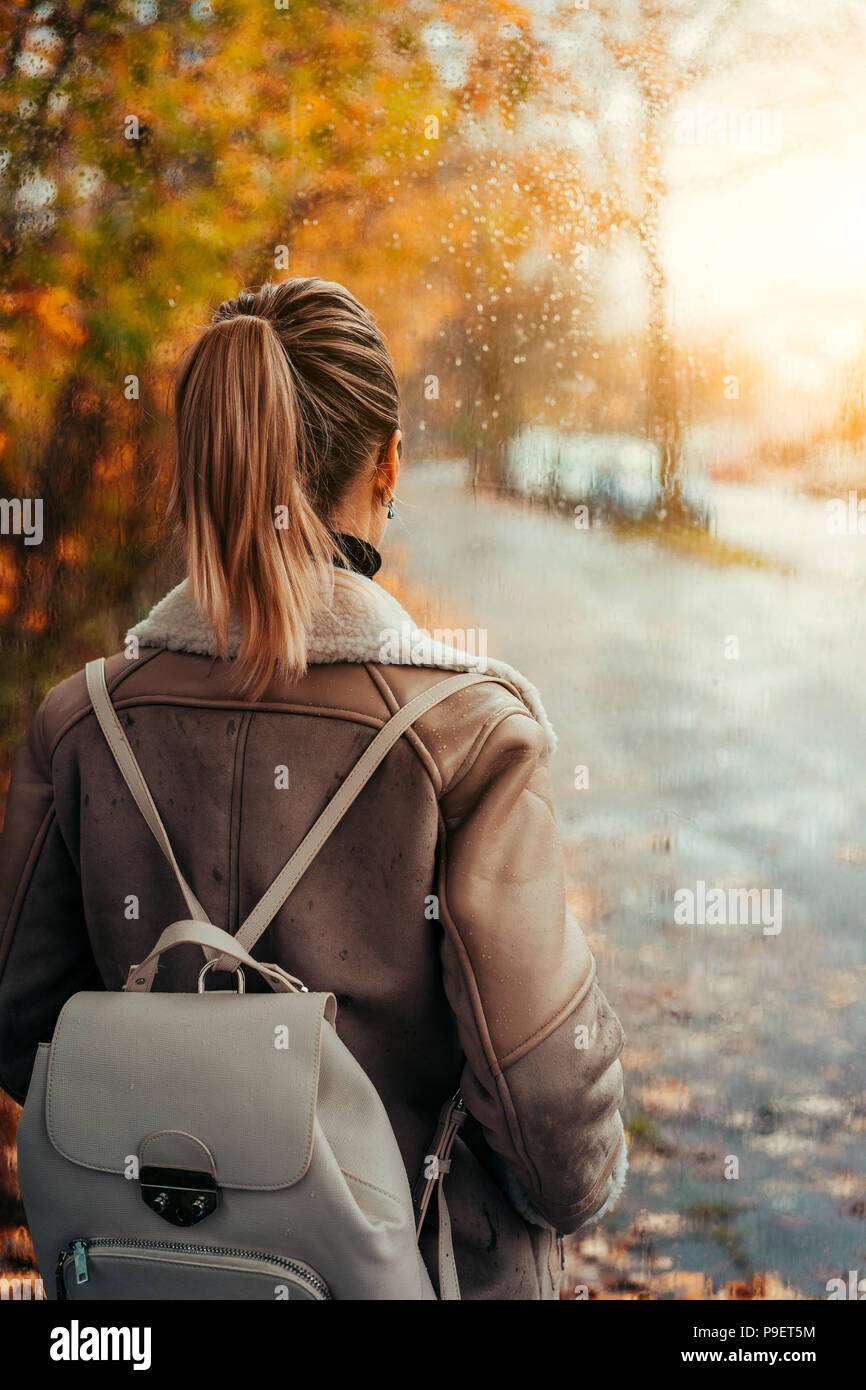 Frau tragen Aviator Jacke suchen durch die Fenster mit Regen fällt. Goldener Herbst Blätter Szene. Unscharf, von Bäumen gesäumten Straße unter Abendsonne Stockfoto