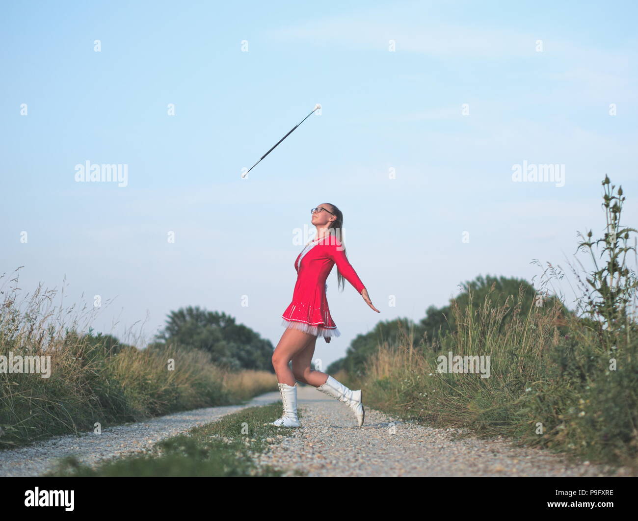 Bebrillter blonder Teen Majorette Mädchen Twirling Baton draußen im roten Kleid Stockfoto