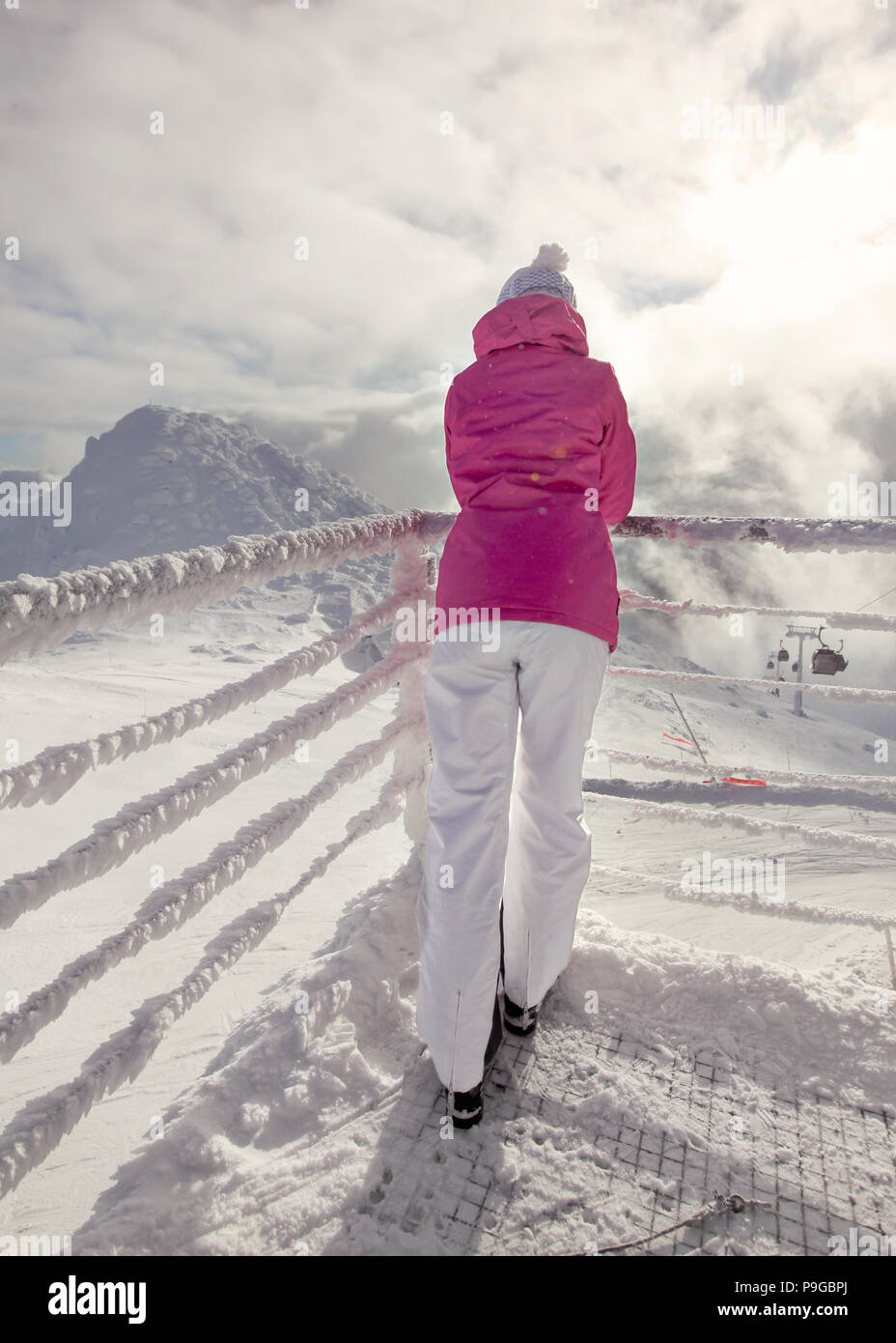 Frau in rosa Winterjacke lehnte sich auf Schnee bedeckten Rampe, Skipisten, mit Sonne und Wolken im Hintergrund Hintergrundbeleuchtung. Stockfoto