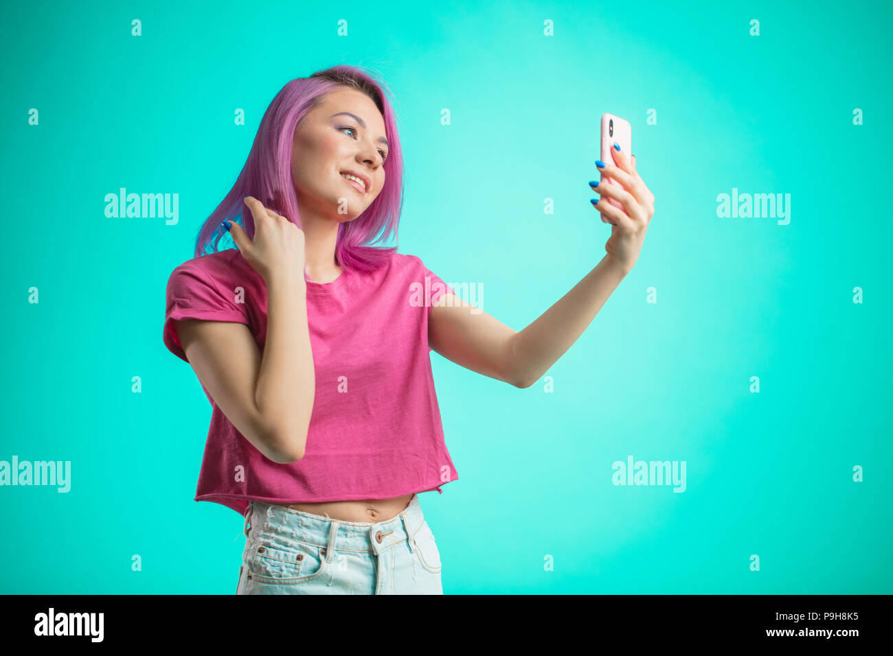 Brustbild mit einem lächelnden Frau mit rosa Haaren, in rosa T-Shirt gekleidet, so dass selfie Foto auf dem Smartphone auf einem blauen Hintergrund mit Stockfoto