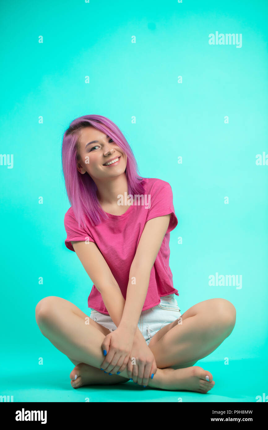 Fröhliches Mädchen mit rosa Haaren Sitzen mit gekreuzten Barfuß leggs am Studio blauen Boden. Stockfoto