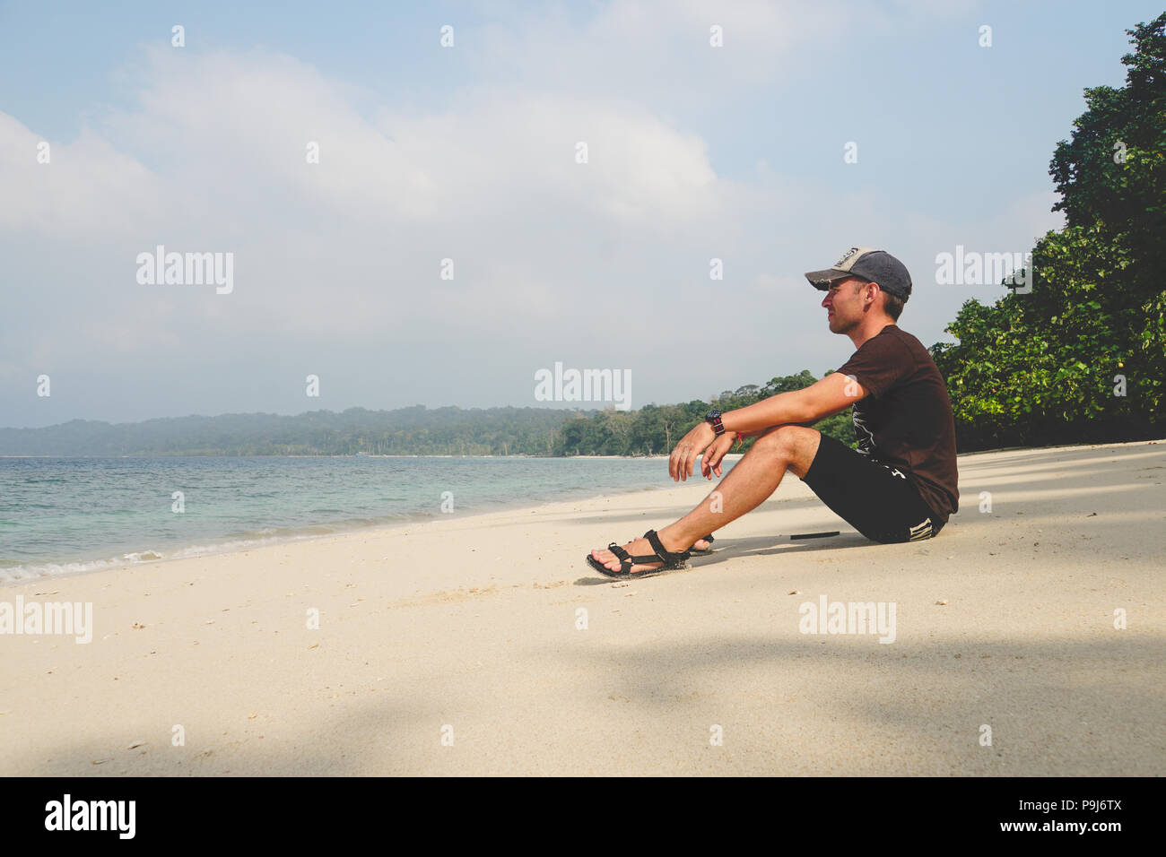 Ein junger Mann sitzt auf einem weißen Sandstrand und schaut auf das Meer auf einer exotischen Insel. Mann auf einem leeren tropisch-exotischen Strand. touristische Besucher reisen Stockfoto