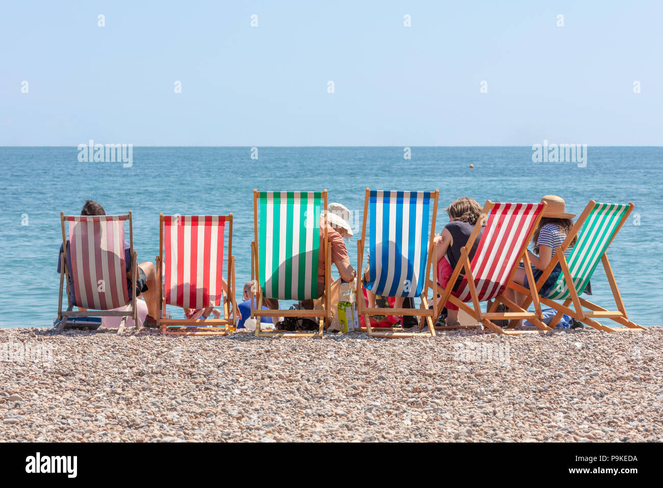 Ansicht der Rückseite des Gruppe von Menschen in sechs Gestreifte Liegestühle am Meer auf einem sonnigen Sommertag sitzt Stockfoto