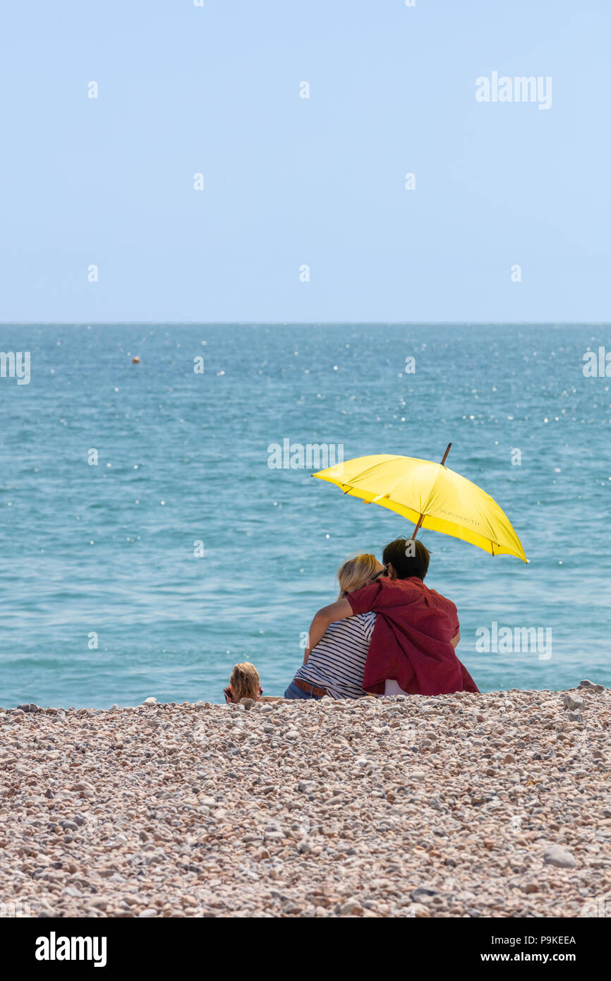 In Worthing, Großbritannien; 14. Juli 2018; Rückansicht Paar am Strand unter einem gelben Regenschirm im heißen Sommer Wetter Stockfoto