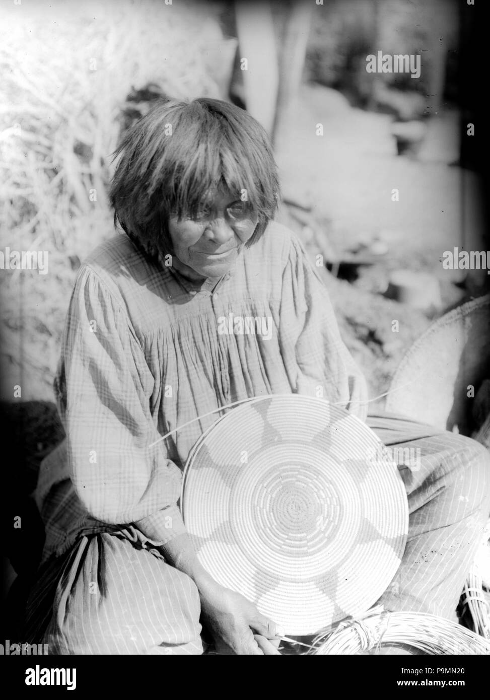 . Englisch: Apache Indian Frau mit einem Korb, ca. 1900 Foto einer Apache Indian Frau mit einem Korb, ca. 1900. Sie sitzt crosslegged auf dem Boden mit einer teilweise bearbeiteten flachen Korb in die Hände in ihren Schoß. Teil eines großen Korb sichtbar ist neben ihr sitzen. Sie trägt ein Plaid Shirt über ein gestreiftes Kleid. Ihr Haar über die Schultern abgeschnitten und hat Bangs. Pinsel wächst hinter ihr. Rufnummer: CHS-3582 Fotograf: James, George Wharton Dateiname: CHS-3582 Abdeckung Datum: ca. 1900 Teil der Sammlung: California Historical Society Collection, 1860-1960 Typ: Bilder Pa Stockfoto