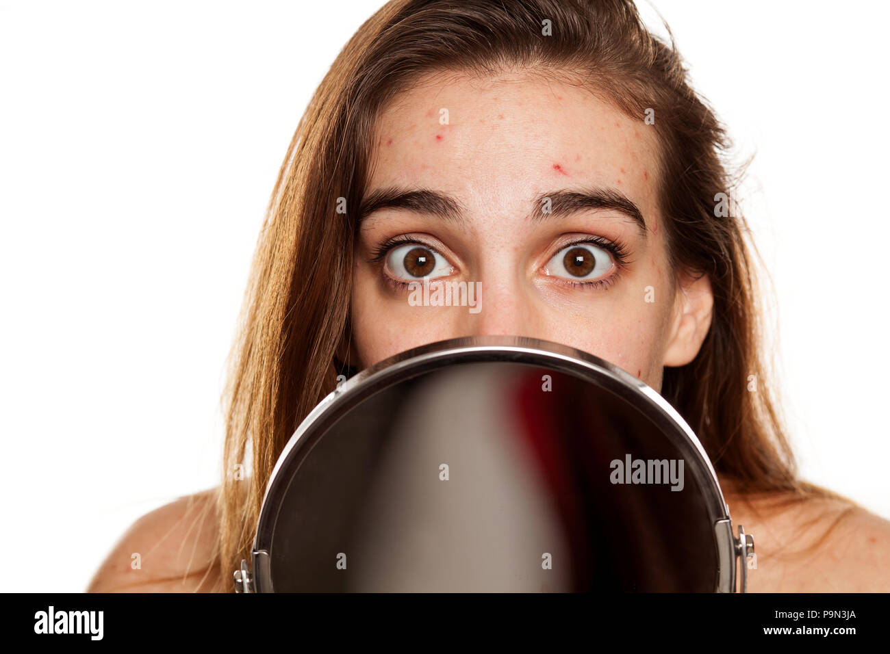 Junge unglückliche Frau mit problematischer Haut und ohne Make-up Spähen hinter dem Spiegel auf weißem Hintergrund Stockfoto