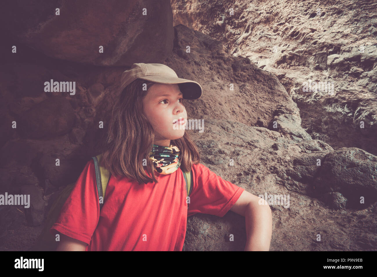 Ein Mädchen starrt nachdenklich in der Mitte einer langen Wanderung, geschützt unter einigen Felsen an einem Berghang. Sie trägt ein UV-Schal, Mütze, Sonnencreme. Stockfoto