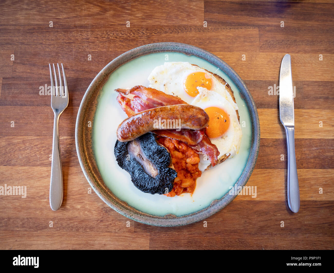 Ein englisches Frühstück mit Speck, Eier, Würstchen, Bohnen und Pilzen auf einem Schild an einem hölzernen Tisch mit Messer und Gabel Stockfoto