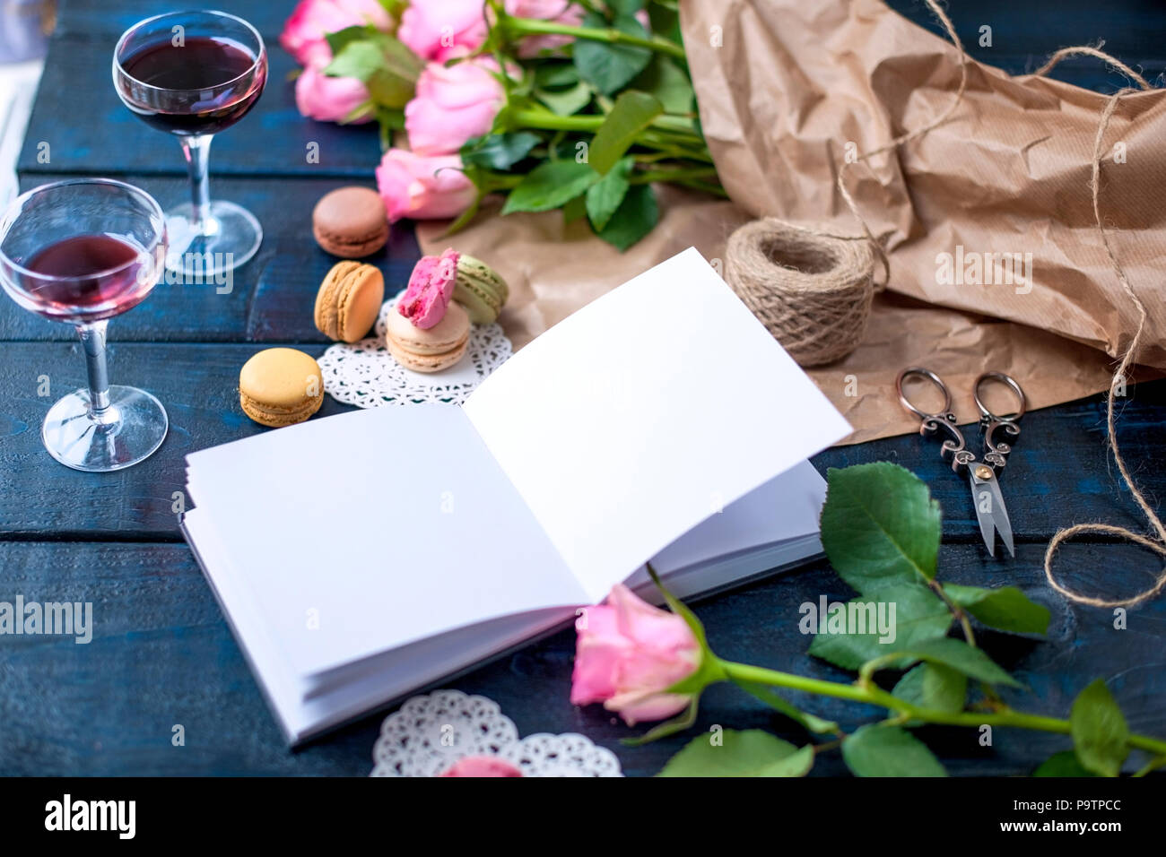 Ein Blumenstrauß aus Rosen ist rosa, in grauem Papier und ein weißes Herz. Notebook öffnen. Zwei Gläser Rotwein. Süße nudeln Makronen in verschiedenen Farben, am Valentinstag. Ein freier Platz für Text oder Werbung. Stockfoto
