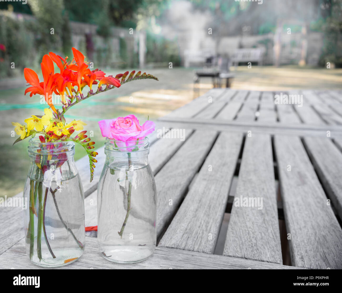 Ein paar Töpfe mit bunten Blumen auf einem hölzernen Tisch in einen englischen Garten an einem sonnigen Sommertag, mit Grill im Hintergrund. Stockfoto