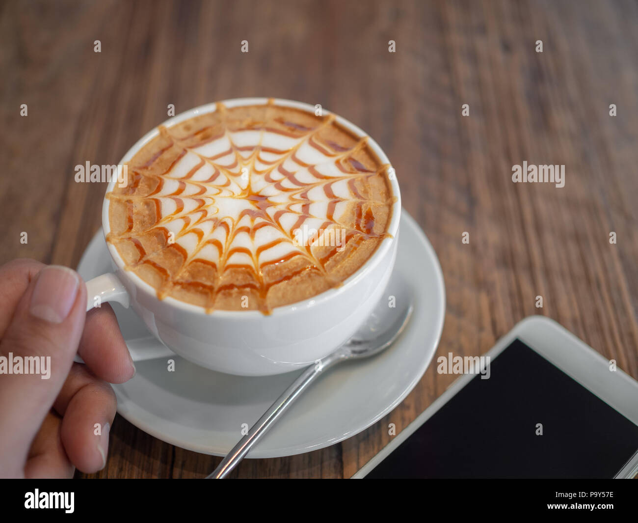 Schone Caramel Macchiato Kunst an der Spitze. HeiBer Kaffee in weiBe Tasse mit silbernen Loffel in der Hand und weiB Handy auf Holz Hintergrund Stockfotografie Alamy
