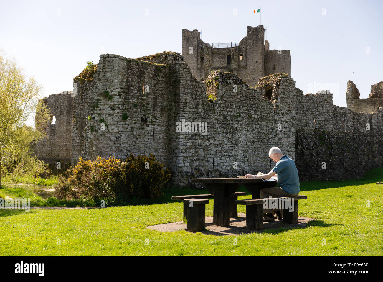 Verkleidung, Irland - Mann sitzt auf einer Picknickbank und lesen Morgen Papier vor einem anglo-normannischen Trim Castle in der Grafschaft Meath. Stockfoto