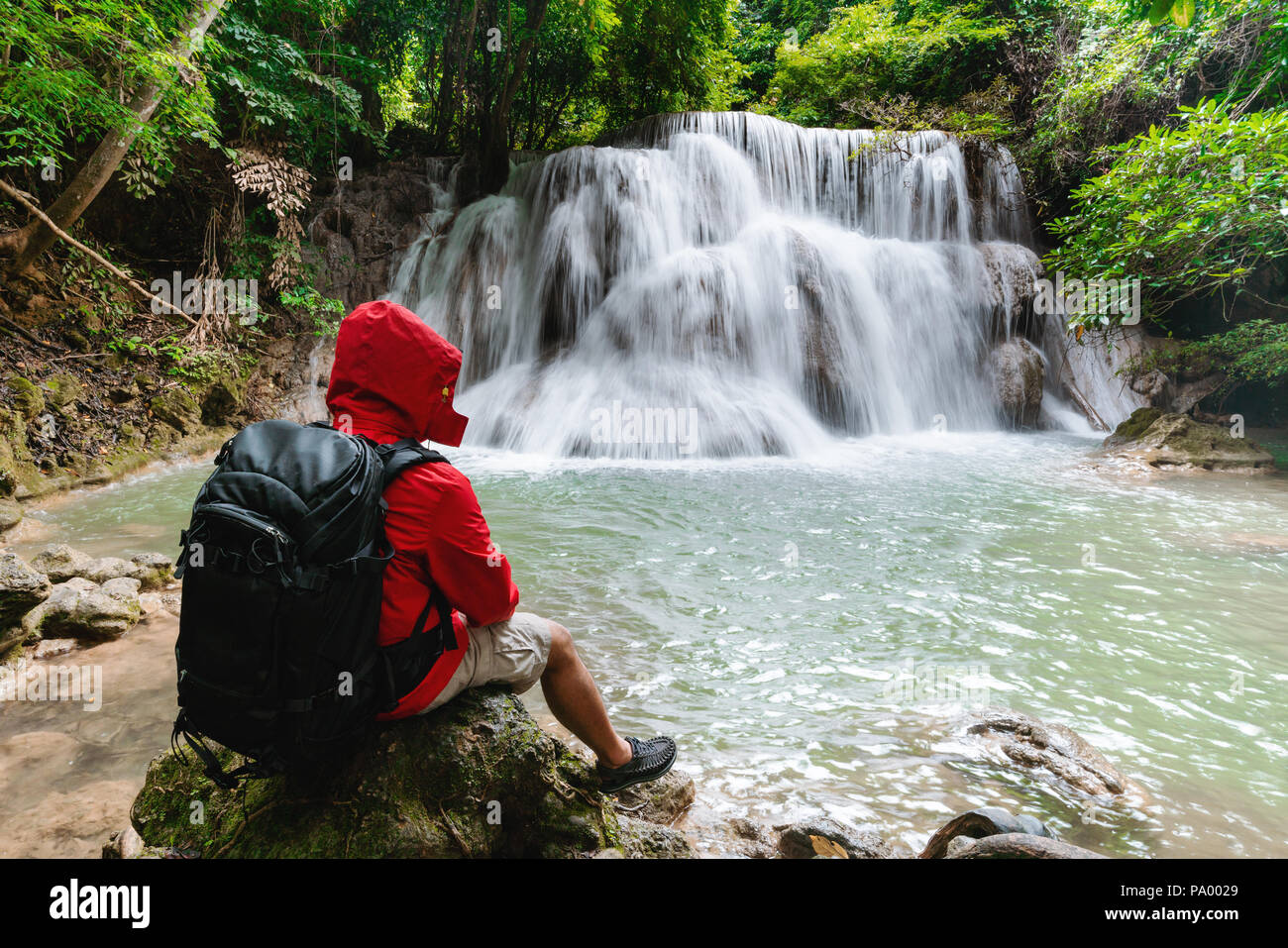 Ein Mann im roten Mantel mit Rucksack auf dem Felsen genießen Sie einen wunderschönen Wasserfall in tropischen Regenwald Huay Mae Khamin in Kanchanaburi, Thailand sitzen Stockfoto