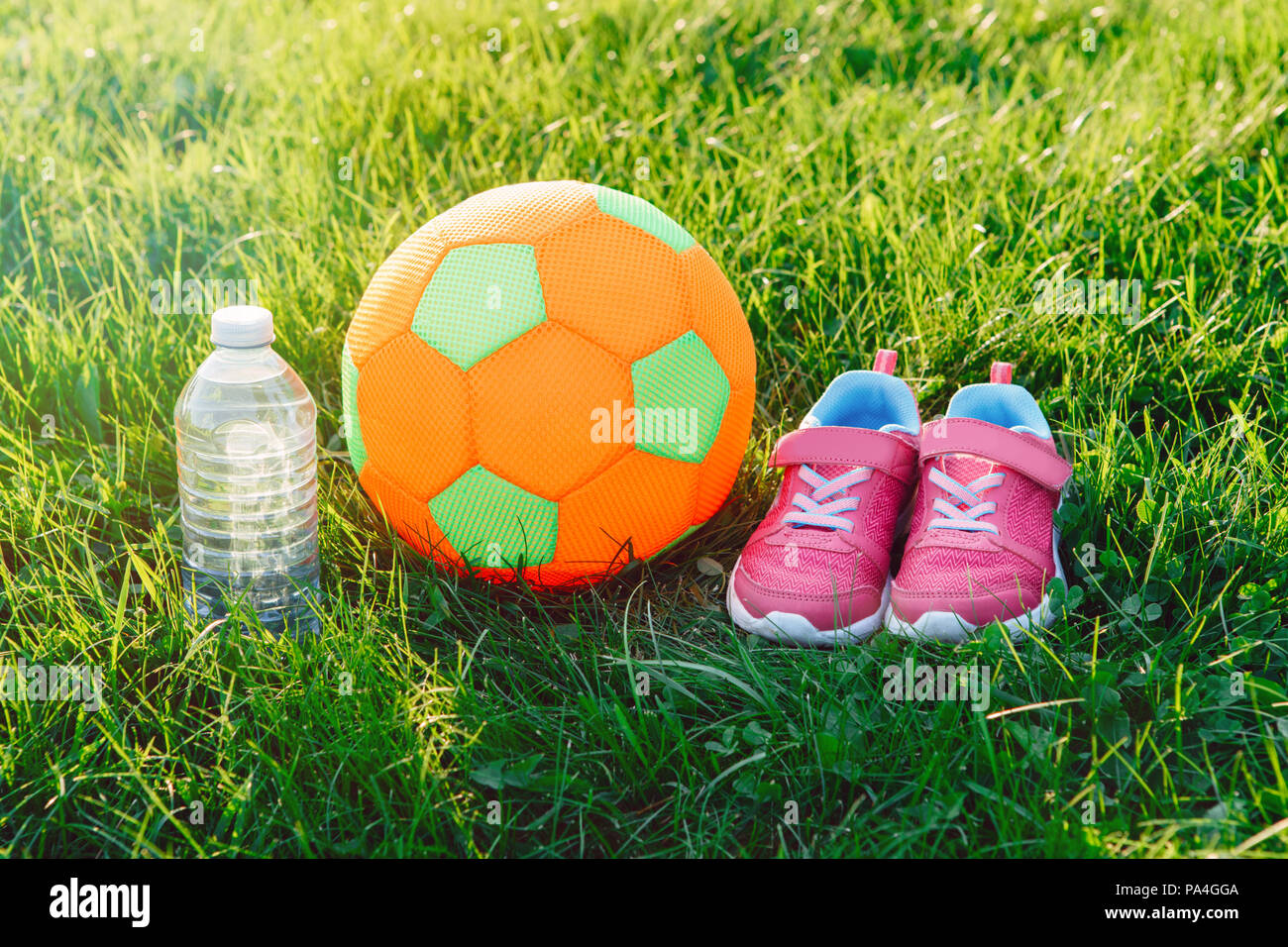 Paar rosa Mädchen Sneaker Schuhe, Kind Stoffverdeck Fußball und eine Flasche Wasser im grünen Gras draussen auf Sonnenuntergang Licht am Abend. Aktivität recreati Stockfoto