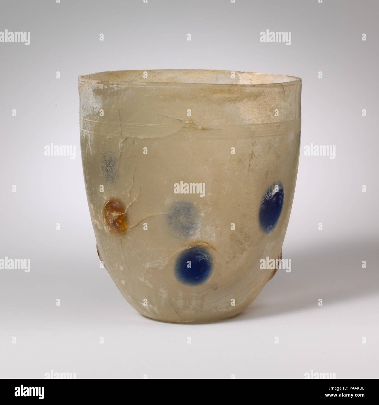 Becherglas. Kultur: Roman, Rheinische. Abmessungen: H.: 4 13/16-in. (12,3 cm) Durchm.: 4 cm. (11,1 cm). Datum: 4.Jahrhundert N.CHR.. Farblos; durchscheinende Kobaltblau und Honiggelb blobs. Uneinheitliche vertikale Rim, geknackt und Boden, leicht outsplayed; hohe, gewölbte Seite zu Körper, verjüngt sich nach unten; konkave Unterseite. Unmittelbar unter Rim, ein einzelnes Rad - horizontale Linie geschnitten; unten, eine Band, bestehend aus zwei Rad - horizontale Linien mit Rad Schnitt - abgenutzte Linien zwischen ihnen, um mittleren und unteren Teil der Seite ein Band der angewandten Blobs, bestehend aus zwei unregelmäßige horizontale Linien von vier wechselnde s Stockfoto