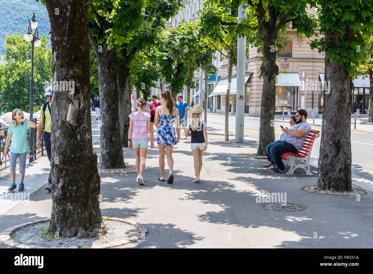 Kleine Stadt Szene, Touristen, Menschen zu Fuß entlang einem Baum Promenade gesäumt, man sitzt auf der bank Telefon, Lugano, Schweiz Stockfoto