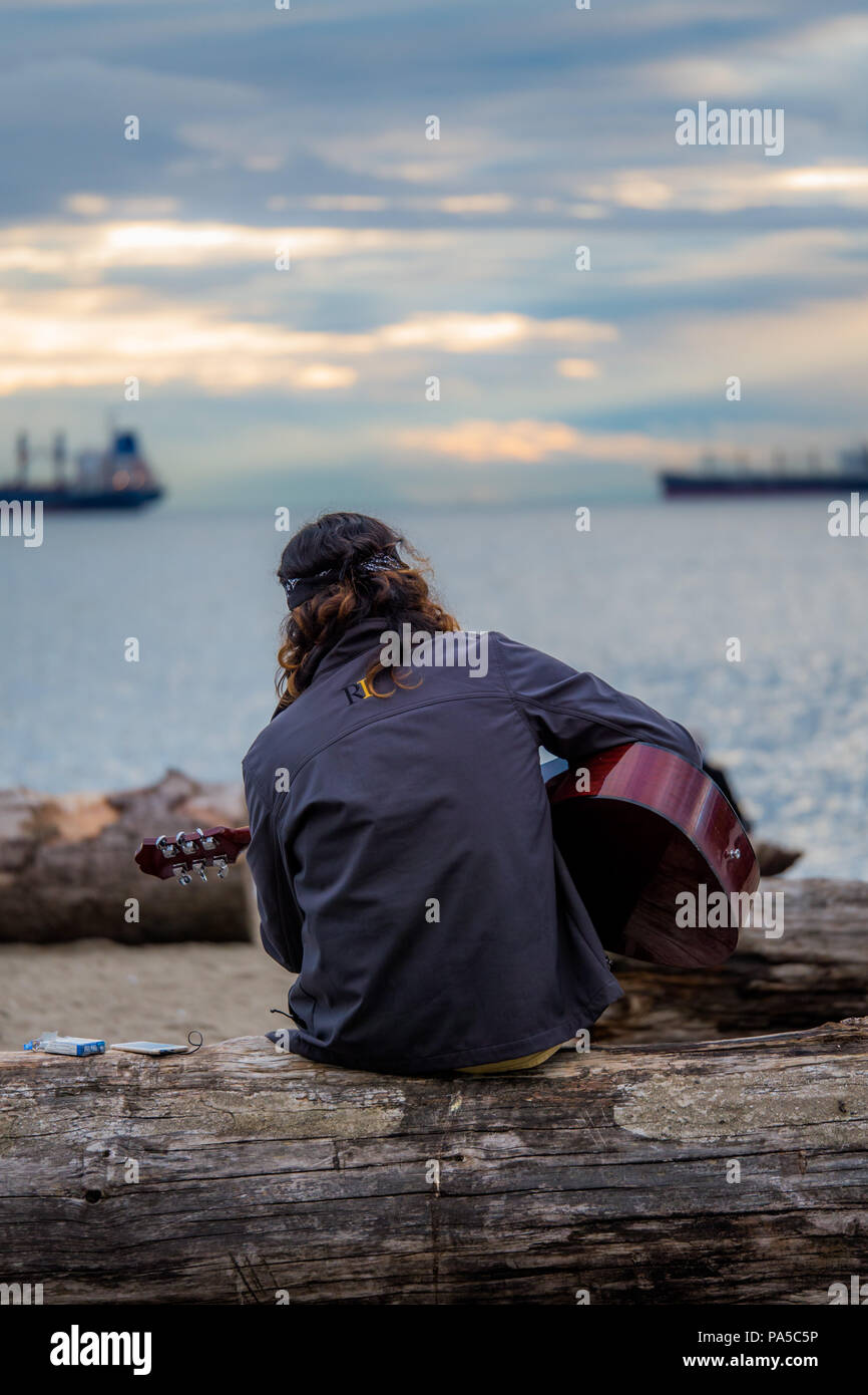 Kopfbügel Guitar Player sitzen auf grob behauenen Log mit Blick auf die Bucht mit der untergehenden Sonne seine Gitarre spielen. Stockfoto