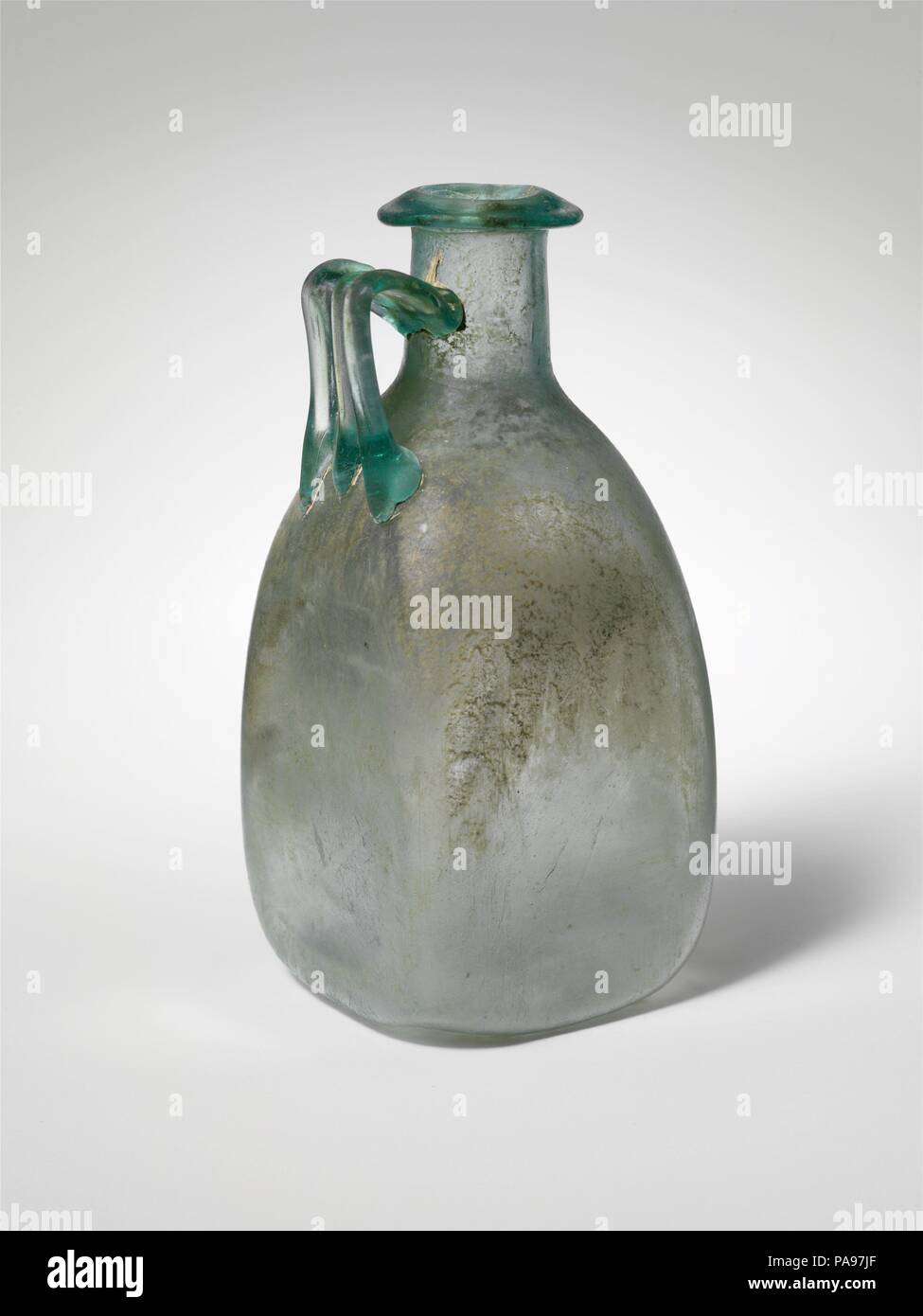 Glaskanne. Kultur: Roman. Abmessungen: Andere: 5 3/4 x 2 5/8 in. (14,6 × 6,7 cm). Datum: 1.-2. Jh. N.CHR.. Transluzent blau grün, mit der gleichen Farbe. Rim ausgeklappt, unten, oben und in den Mund, mit der abgeschrägten Oberfläche; zylindrische Hals, leicht nach unten erweitert, mit leichten Schnittspuren um die Basis; schräge Schulter mit abgerundeten Ecken; quadratische Körper mit leicht konkaven Seiten; ungleichmäßige, konkav unten; Strap Griff mit drei Rippen, auf der Schulter, senkrecht nach oben gezeichnet, dann gebogen und ab und an den Hals mit Rückwärts über Trail befestigt. Intakt, außer einem sm Stockfoto