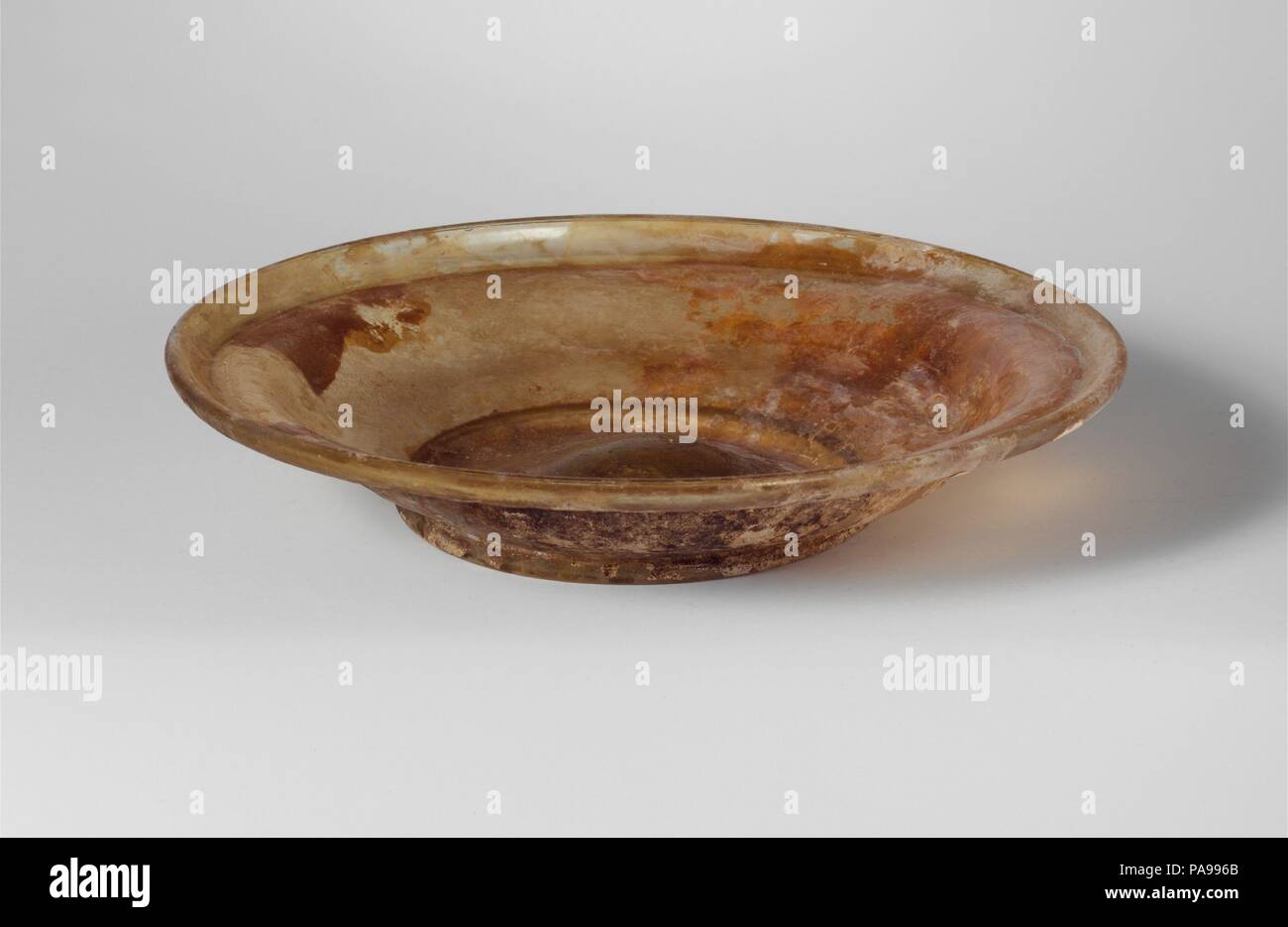 Glas Schale. Kultur: Roman. Abmessungen: Höhe: 1 cm. (3.2 cm) Durchm.: 6 1/4 x 3 1/2 in. (15,9 x 8,9 cm). Datum: 4.Jahrhundert N.CHR.. Farblos mit gelben Farbton. Röhrenförmig, abfackeln Rim, Ausgeklappt, unten, und in den, mit schmalen, leicht konvexen inneren Schulter; S-förmige Seite zu Körper, in geschwungenen zur Integral Tubular Base Ring; Dreschtrommel unten und in der Mitte verdickt und Spuren von kleinen pontil markieren. Intakt; pinprick blasen; Lochfraß, Abstumpfung und Schillern, mit die meisten Außen in starkem Zahnschmelz bedeckt - wie Verwitterung. Museum: Metropolitan Museum of Art, New York, USA. Stockfoto