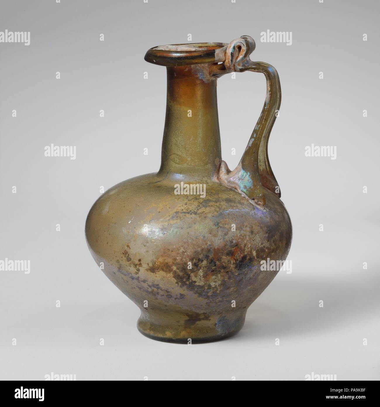 Glaskanne. Kultur: Roman. Abmessungen: 4 5/16 in. (11 cm) Andere: 3. 7,6 cm (3 Zoll) Durchm. Von rim: 1 5/16 in. (3.3 cm) Durchm. Der Fuß: 1 9/16-in. (3,9 cm). Datum: 1. Jh. N.CHR.. Kleine ein- Kanne Translucent gelb grün; in gleicher Farbe. Rim ausgeklappt, unten, rund, und oben, mit abgeschrägter äußerer Kante über dem Abfackeln Mund, leicht konisch zylindrischer Hals, mit schnittspuren an der Basis; Hocke, bauchigen Körper, in geschwungenen, integral, hohen Basis ring; konkave Unterseite; Strap Griff in zwei breiten Klauen an der vorderen und an der dreieckigen Pad an der Rückseite Ausbreitung auf der Unterseite des Stutzens mit Werkzeug zu Schulter Stockfoto