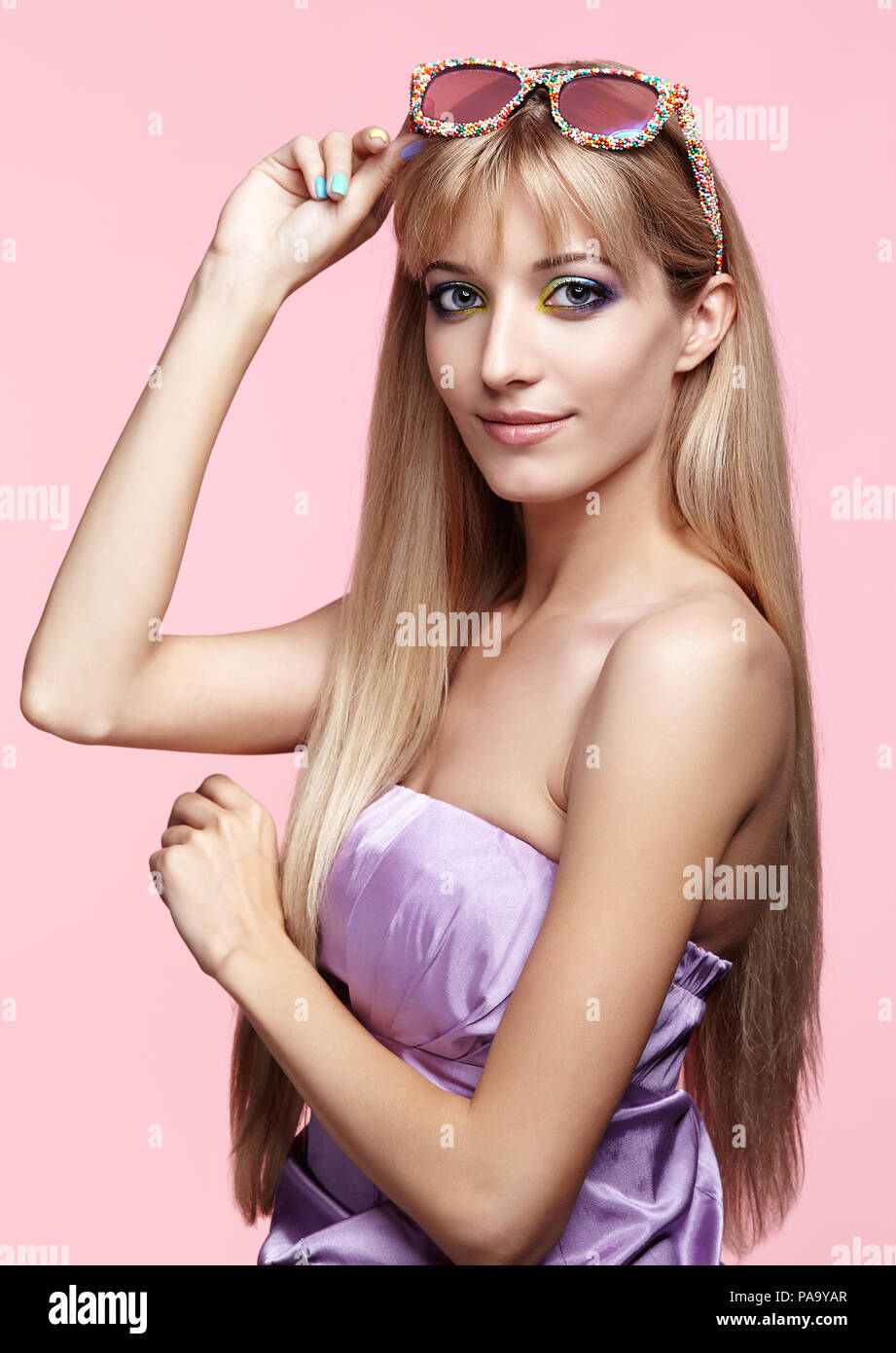 Blonde junge lächelnde Frau mit Spaß Süßigkeiten Gläser auf rosa Hintergrund. Porträt von Frauen in lila Kleid mit blauen und gelben Augen Make-up. Stockfoto
