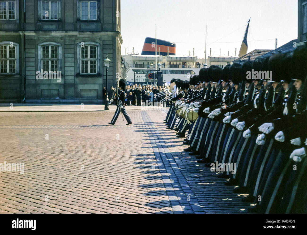 Eine Menschenmenge, die die dänische Kaisergarde in Uniformen und Bärenfell-Hüten beim Wachwechsel im Schloss Amalienborg in Kopenhagen in den 1960er Jahren beobachtet. Der Fährhafen von Oslo ist an der Stelle sichtbar, an der sich der Amalie Park befindet Stockfoto