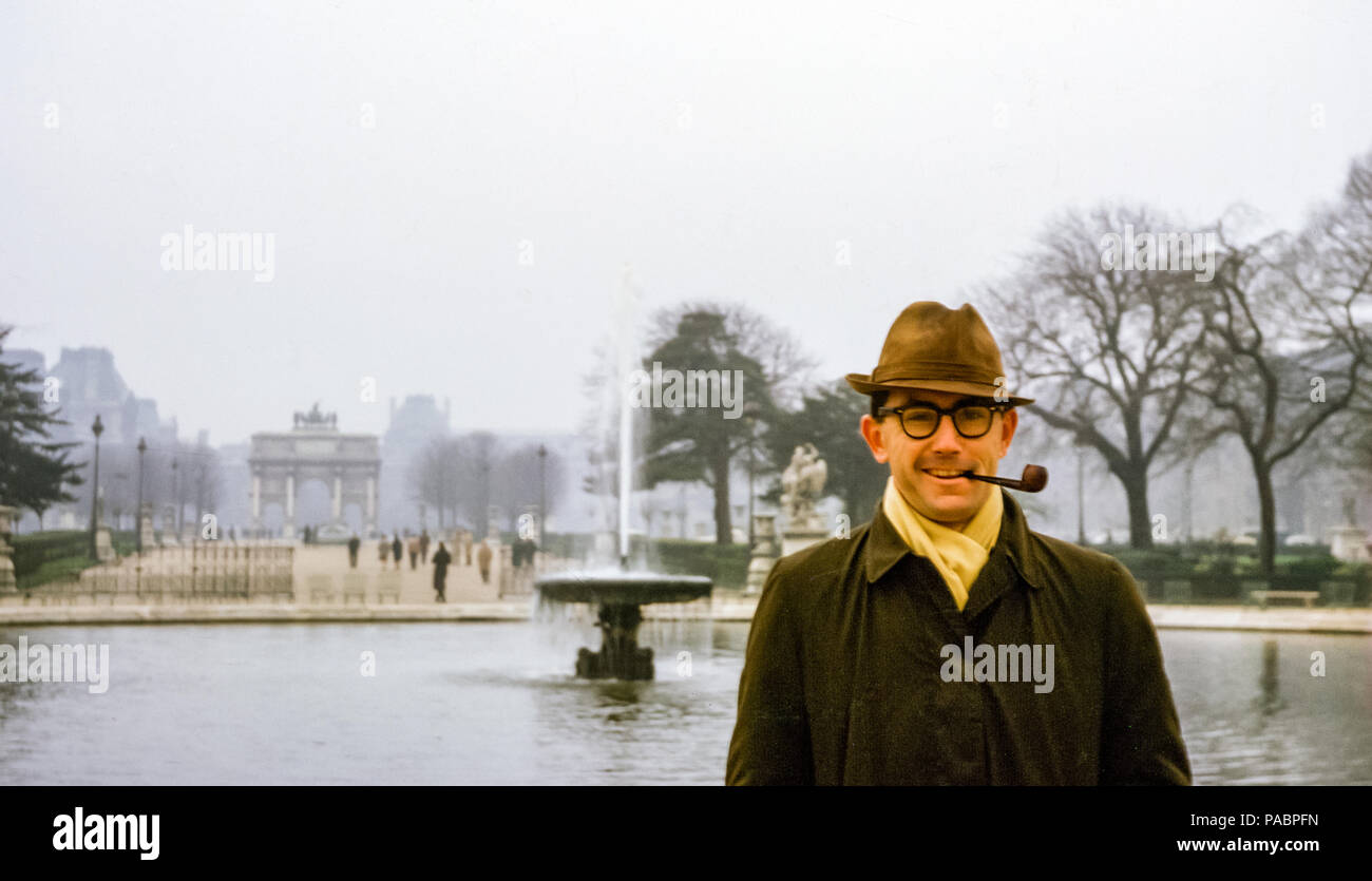 Amerikanischer Tourist mit Trilby-Hut, der eine Pfeife raucht und vor einem Brunnen auf dem Place du Carrousel steht, mit dem Arc de Triomphe in den 1960er Jahren Stockfoto