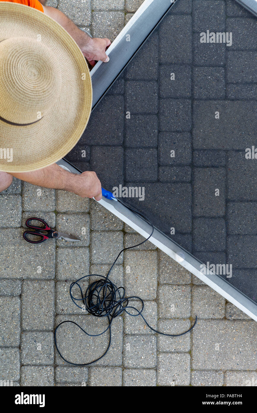 Mann Reparatur eines beschädigten Bildschirm Tür auf eine Terrasse nach einem Sturm der Anbringung der neuen Wire Mesh in den Metallrahmen von oben tragen ein Stroh sonnenhut gesehen Stockfoto