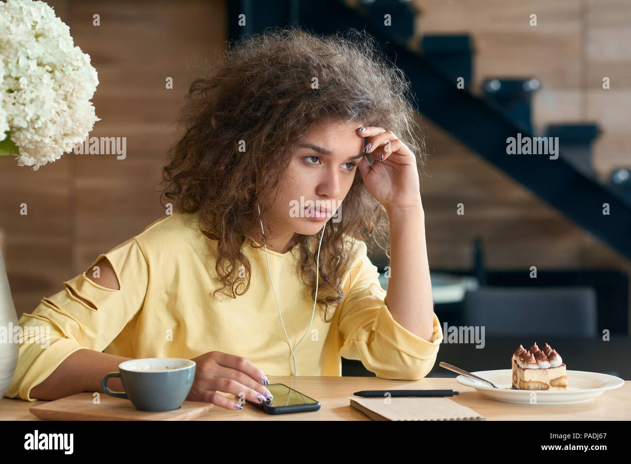 Deprimiert junge Frau mit lockigem Haar sitzt auf dem Tisch des Holz- Restaurant, erschöpft und wütend, Gefühl negative Emotionen. Tasse Kaffee und weiß lila Blumen, Treppen für den Hintergrund. Stockfoto