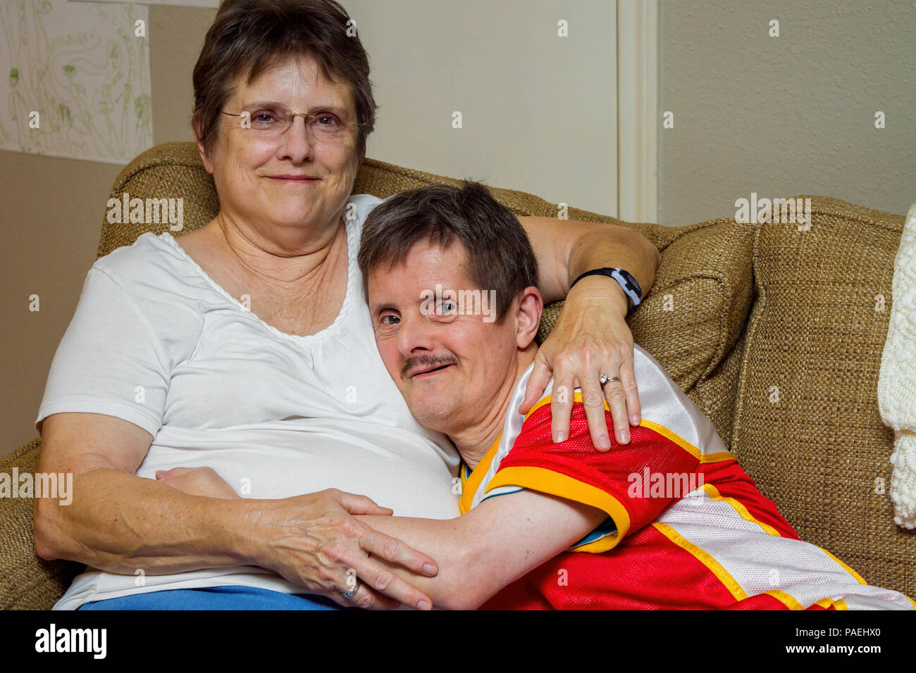 Ein älterer Mann mit Down-syndrom, umarmt seine ältere Schwester, als sie auf der Couch sitzen. Die Frau sieht veraltet, der Mann sieht mischeivious. Er ist, zu tic Stockfoto