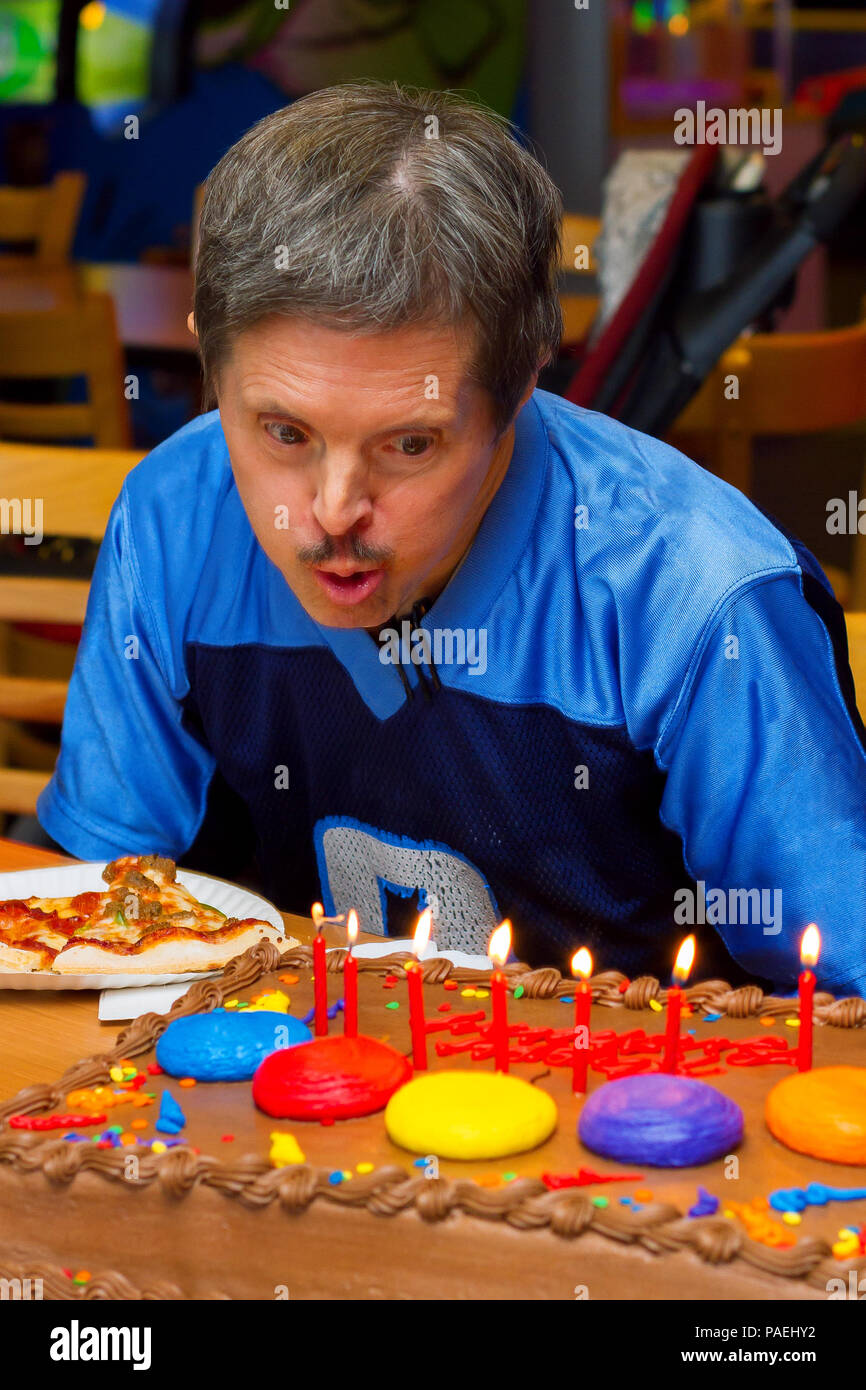 Ein älterer Mann mit Down-syndrom bläst die Kerzen auf dem Geburtstagskuchen an einer Pizza Party in seiner Ehre. Er sieht sehr konzentriert, wie er versucht zu löschen. Stockfoto