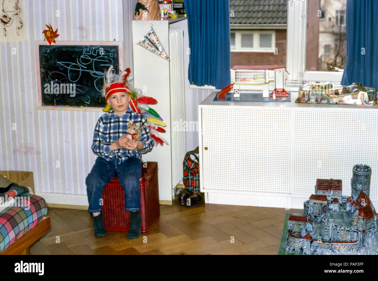 Der Junge trägt einen indianischen Kopfschmuck und sitzt in einem Kinderzimmer mit einem Kuscheltier mit Spielzeug, darunter eine Texaco-Garage und ein deutsches Elastolin-Hausser-Plastikspielzeug mittelalterliche Burg Schaffhausen Knights Castle, London, England, Großbritannien in den 1960er Jahren Stockfoto