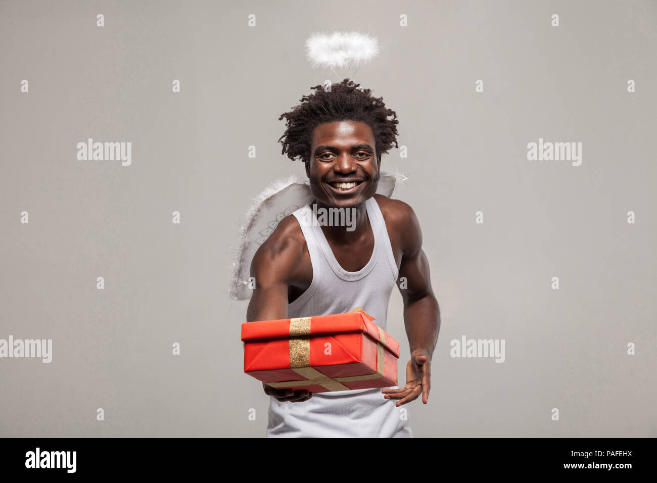 Liebe Konzept und gemeinsame goft. Dunkelhäutige bärtigen African Angel lustiger Mann mit weißem Hemd, Nimbus und Winks atanding und Betrieb vorhanden Geschenkbox. Stockfoto