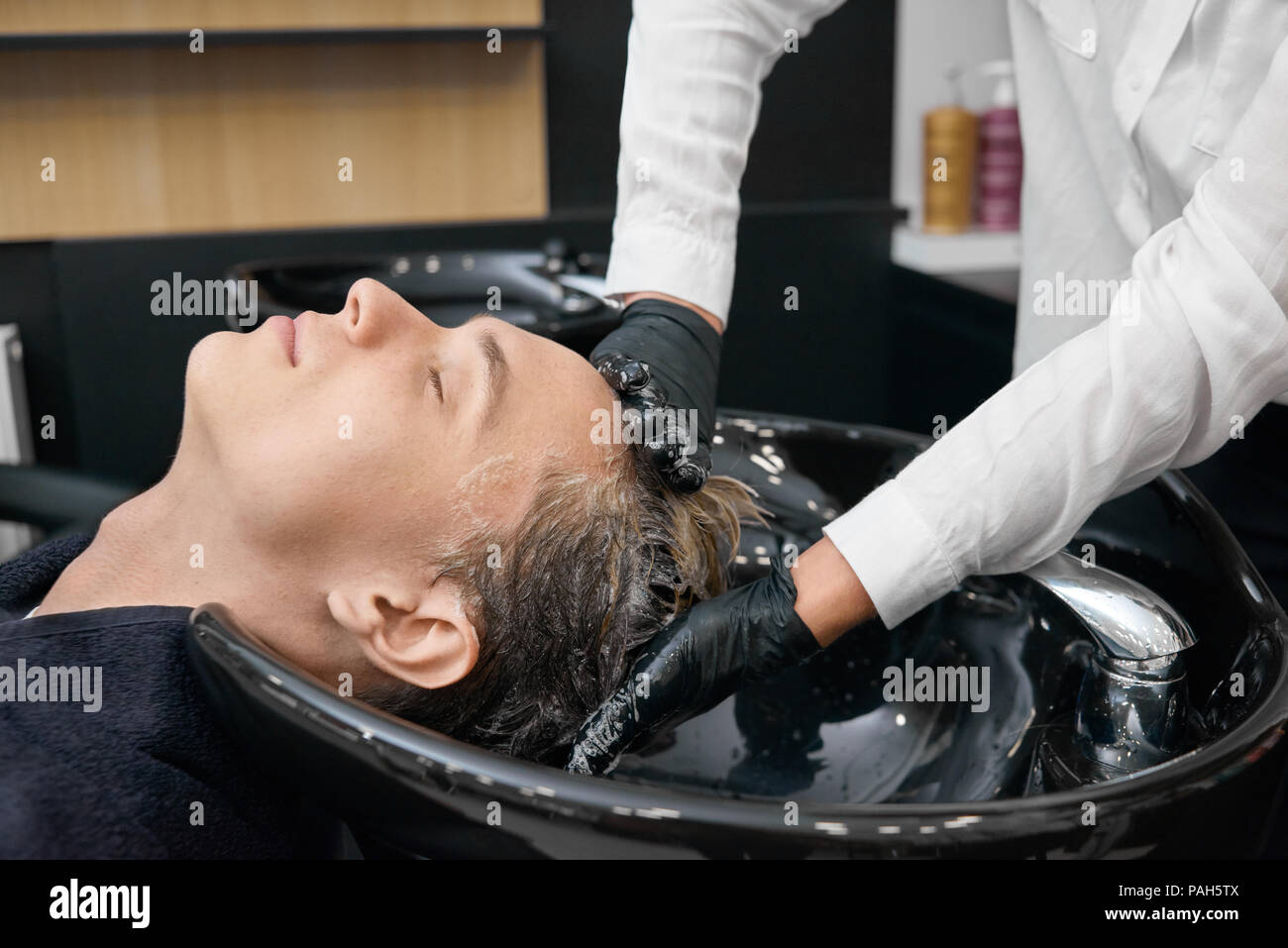 Der Hairstylist waschen client Haare im Waschbecken ein Beauty Salon. Client sitzt mit seinem Kopf in die schwarze Keramik Waschbecken mit geschlossenen Augen. Wohlfühlen, Relaxen. Friseur mit schwarzen Handschuhen. Stockfoto