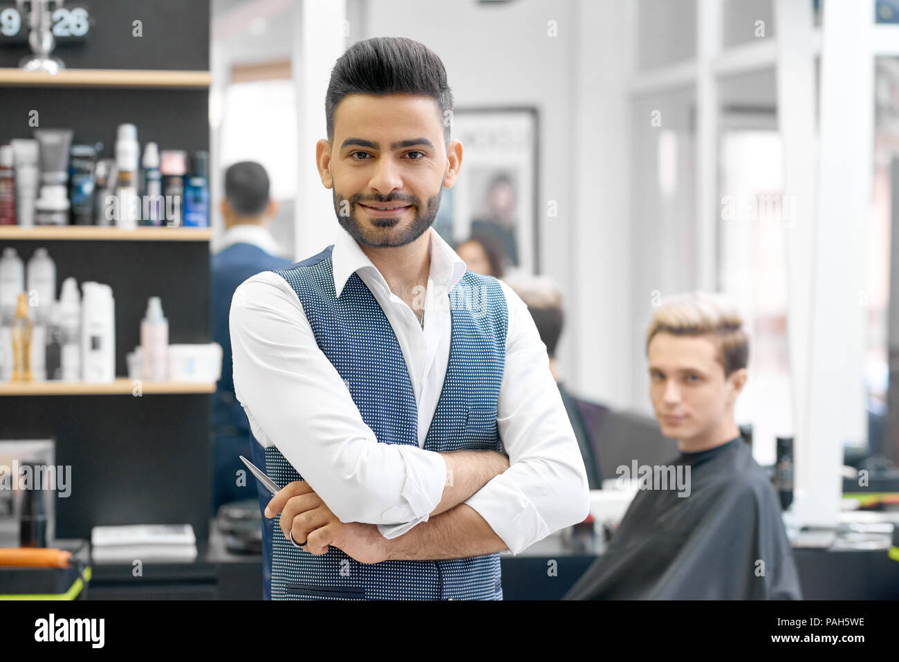 Stattliche friseur Posieren vor der jungen Kunden in der Nähe von Großer Spiegel sitzen. Hairstylist tragen weiße Weste casual Shirt, grau metallic, scharfe Schere, an der Kamera schaut. Stockfoto
