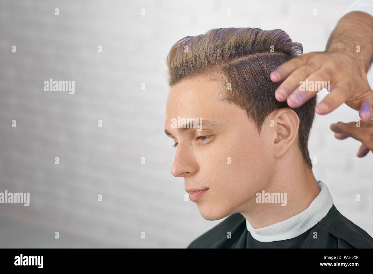 Junge männliche Modell warten auf neue Frisur mit lila Haare färben. Friseur Hände tun, stilvolle Farbgebung für schöner Junge. Arbeiten am weißen Stein studio Hintergrund. Stockfoto