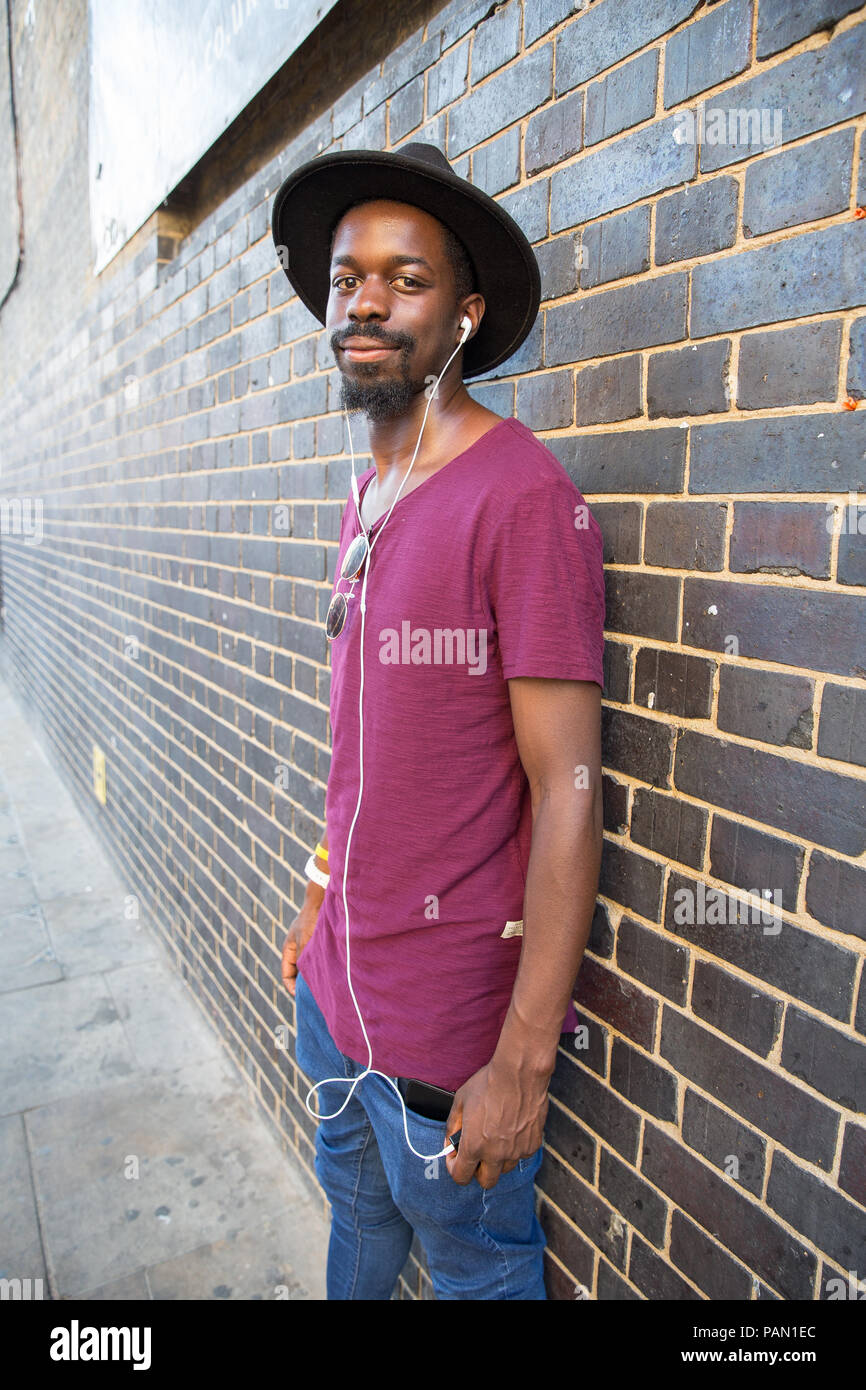 Eine coole Hipster, schwarzer Mann mit dem breiten Hut Krempe, Lila T-Shirt und Ohrhörer, gegen eine Mauer in der Brick Lane, Shoreditch, London lehnend. Stockfoto
