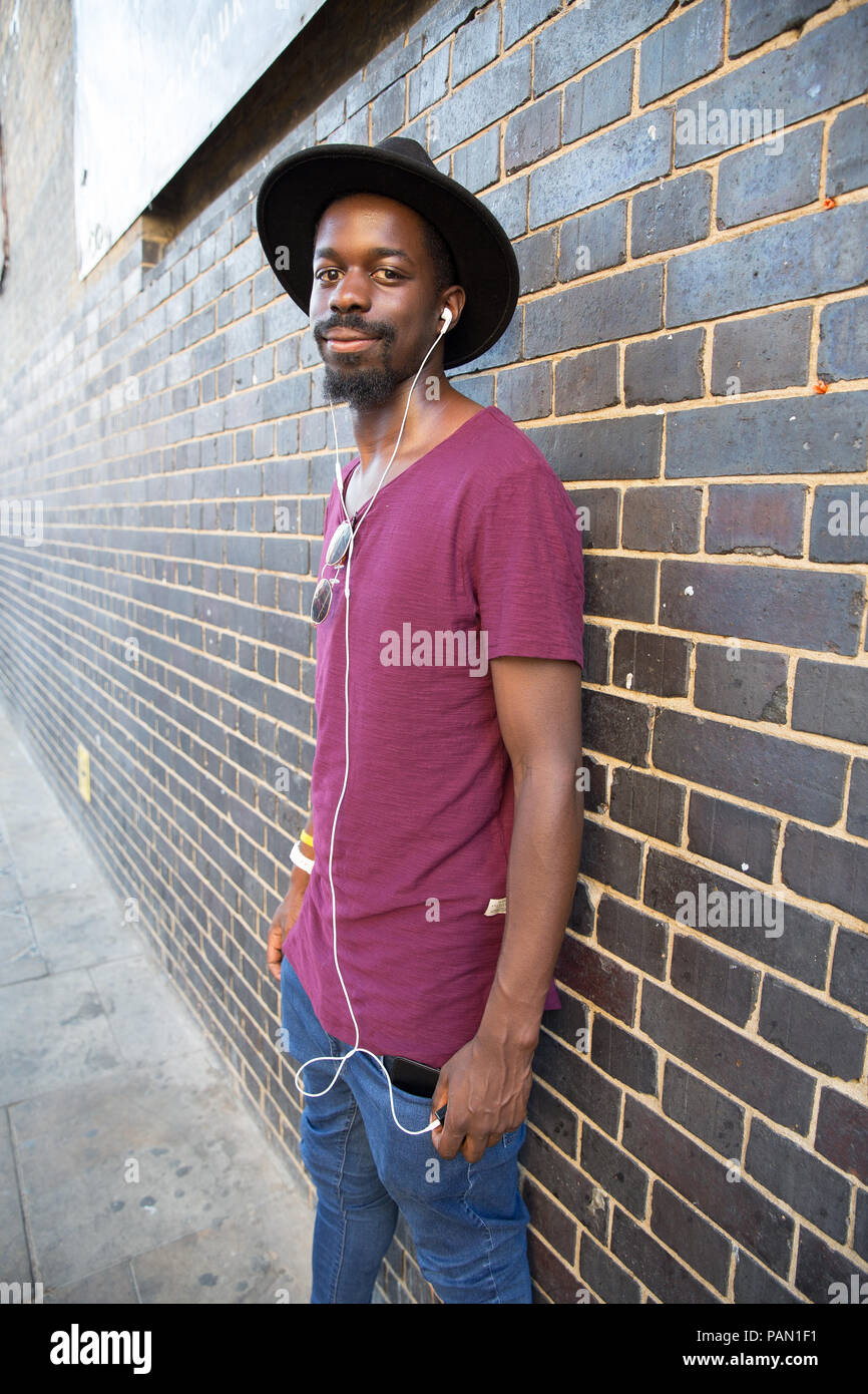 Eine coole Hipster, schwarzer Mann mit dem breiten Hut Krempe, Lila T-Shirt und Ohrhörer, gegen eine Mauer in der Brick Lane, Shoreditch, London lehnend. Stockfoto