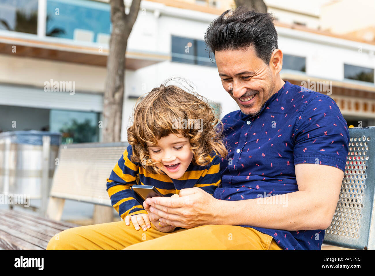 Spanien, Barcelona, glücklich, Vater und Sohn mit einem Smartphone sitzt auf der Bank Stockfoto