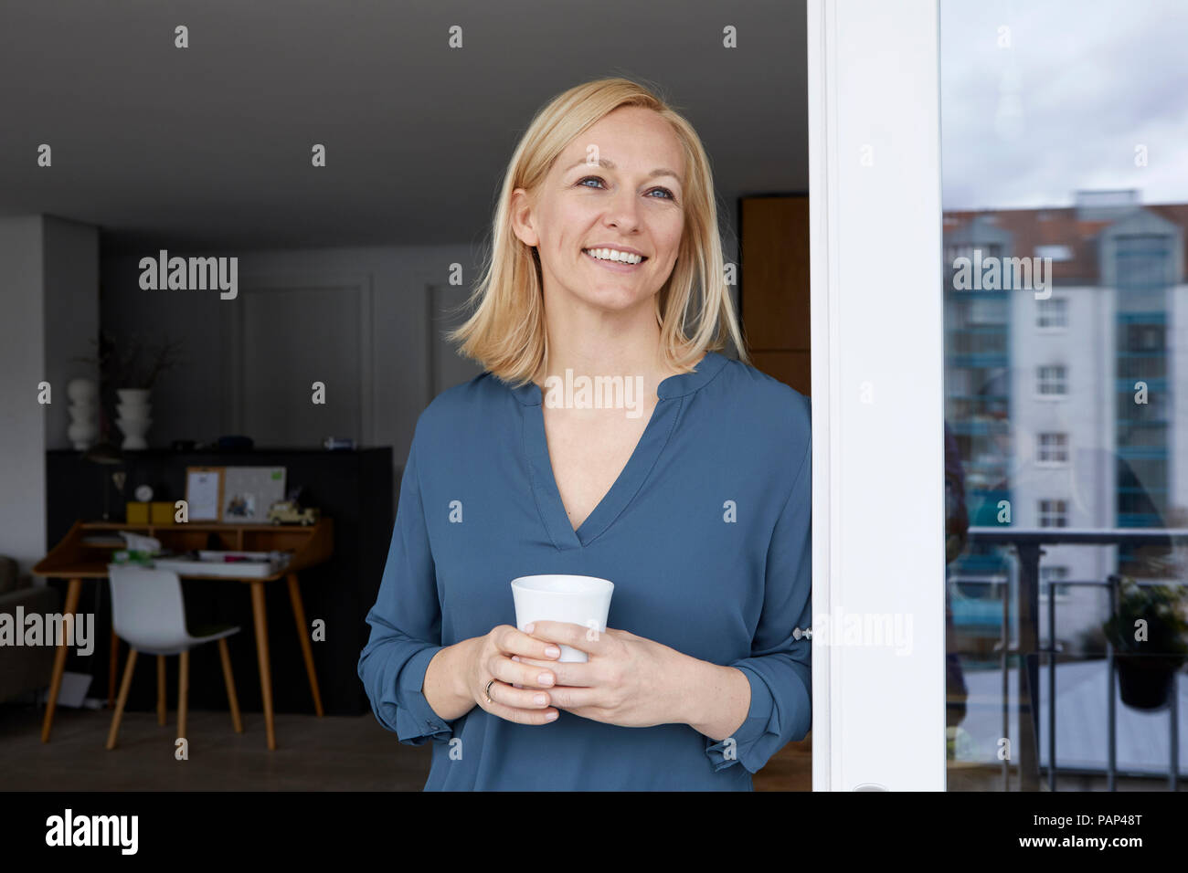Lächelnde Frau mit Tasse Kaffee aus Balkon Tür Stockfoto
