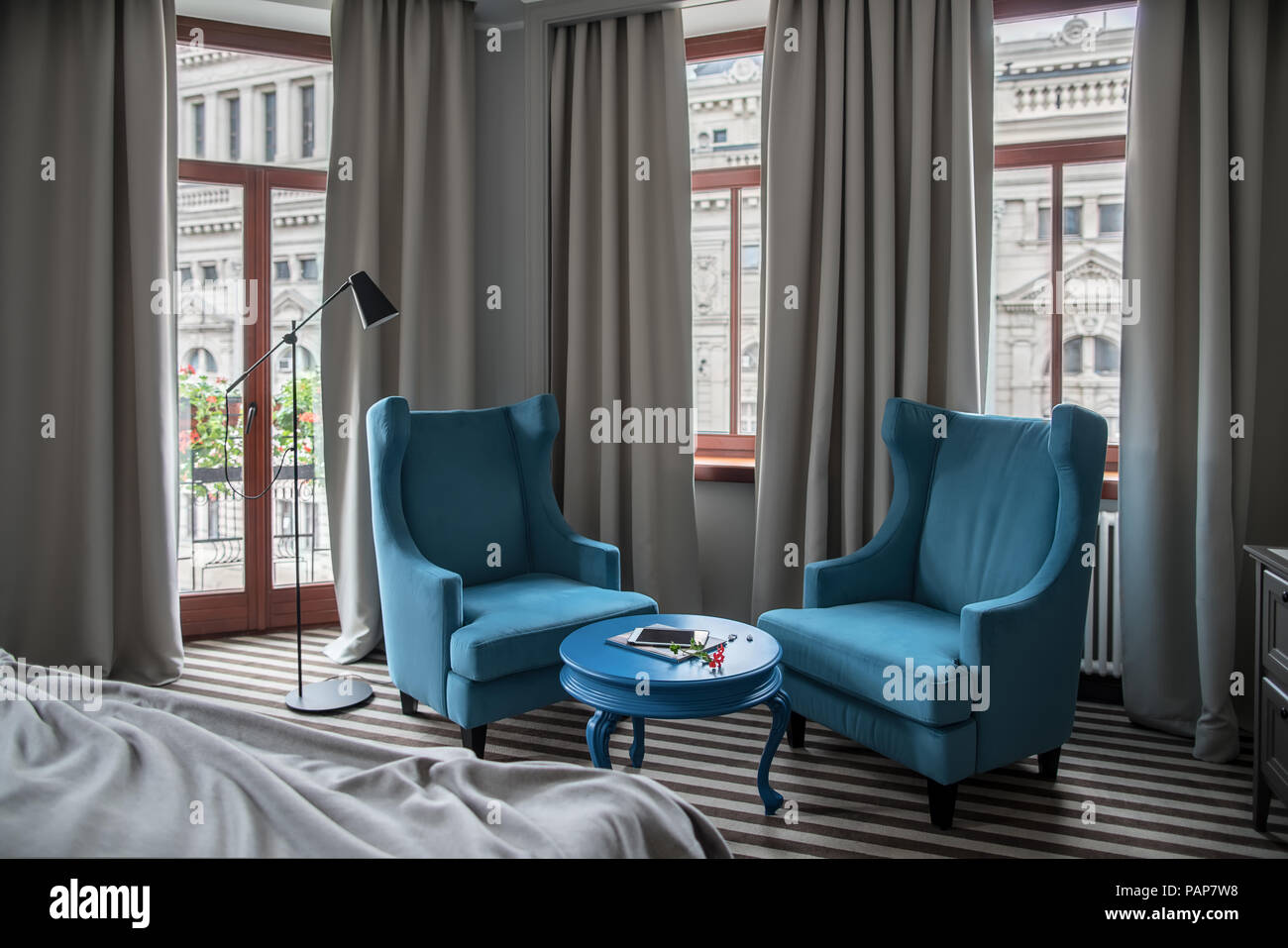 Hotel Zimmer mit hellen Wänden und einem gestreiften Stock. Es ist ein runder blauer Tisch mit zwei Sesseln, Fenster mit Vorhängen, Tür zum Balkon, dunkle Floo Stockfoto