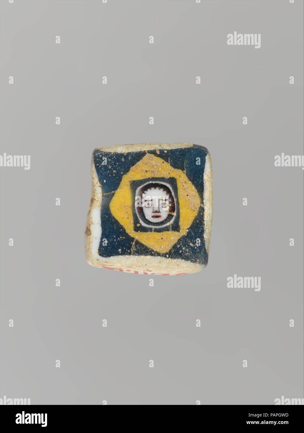 Glas Mosaik Gesicht Raupe. Kultur: Roman, im östlichen Mittelmeer. Abmessungen: H.: 1/2 in. (1,3 cm). Datum: 1. Jahrhundert v. Chr. - 1. nachchristlichen Jahrhundert. Opak Weiß, Gelb und Rot, und translucent Blue und Deep Purple, erscheint schwarz. Platz mit leicht abgerundeten Kanten und Ecken; flach vorne und hinten; kleine horizontale Bohrung. In der Mitte, runde Gesicht von schwarz-weiß gestreiften Haar umrahmt, mit mandelförmigen Augen, Augenbrauen, Nase, Mund und Schlitz mit roten Lippen; das Gesicht in Schwarz und Weiß Kreisen auf ein blaues Quadrat Boden eingeschlossen; dieses wiederum ist in die gelbe Raute auf ein blaues Quadrat, ummantelt Stockfoto