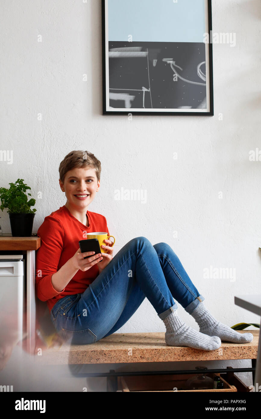 Frau sitzt in der Küche, trinken Kaffee und Kontrolle smartphone Nachrichten Stockfoto