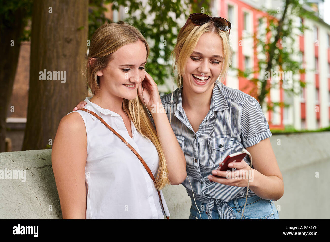Zwei glückliche junge Frauen teilen Handy- und Ohrhörer im Freien Stockfoto