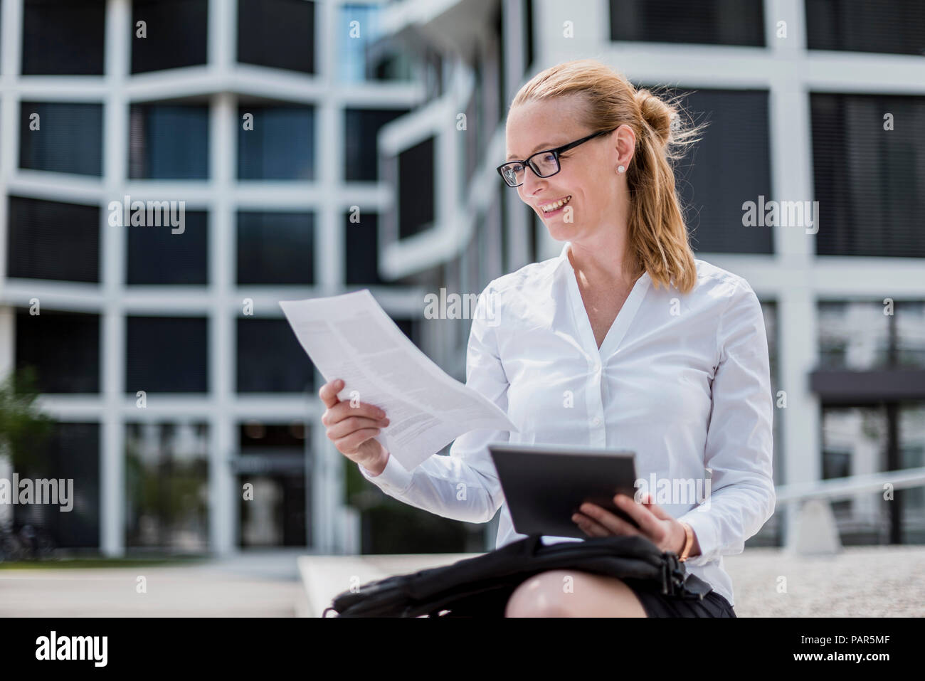 Portrait von lächelnden Geschäftsfrau mit Dokumenten und Tablet vor dem Bürogebäude sitzen Stockfoto