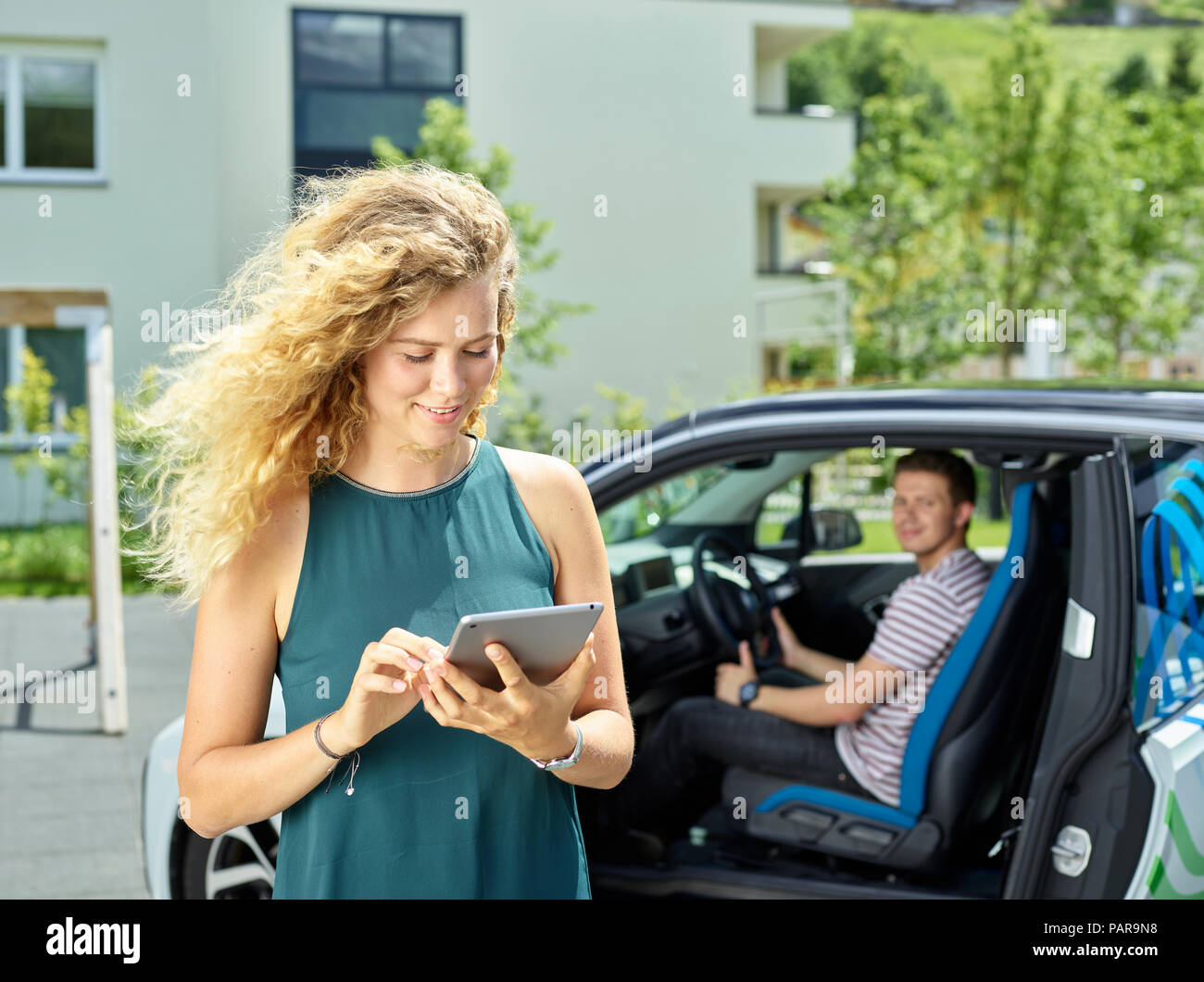 Lächelnde junge Frau mit Tablet mit Mann in elektrischen Auto im Hintergrund sitzen Stockfoto