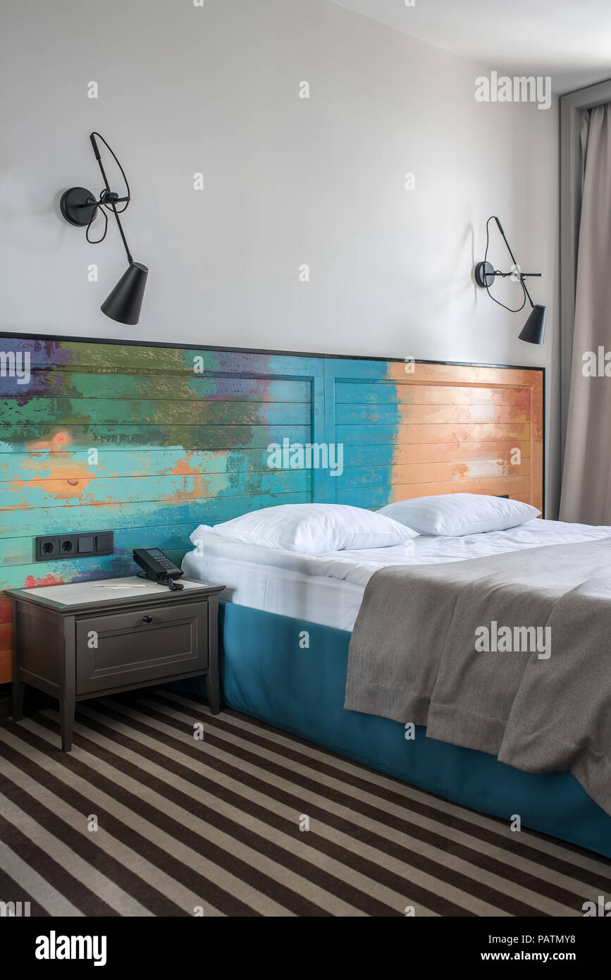 Stilvolle Zimmer in einem Hotel, mit hellen Wänden und gestreiften Stock. Es ist ein blauer Doppelbett mit einem bunten hölzernen bedhead, dunkelgrau Nachttisch mit Pho Stockfoto
