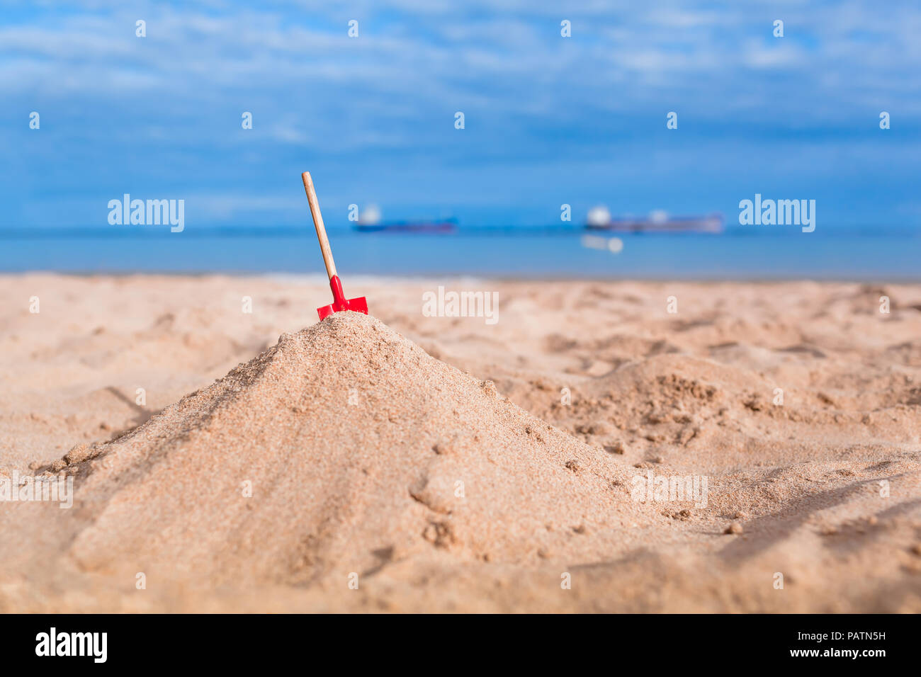 Holiday Beach noch leben, rot Miniatur Schaufel oben Haufen Sand, cargo Schiff auf See Horizont Hintergrund Stockfoto