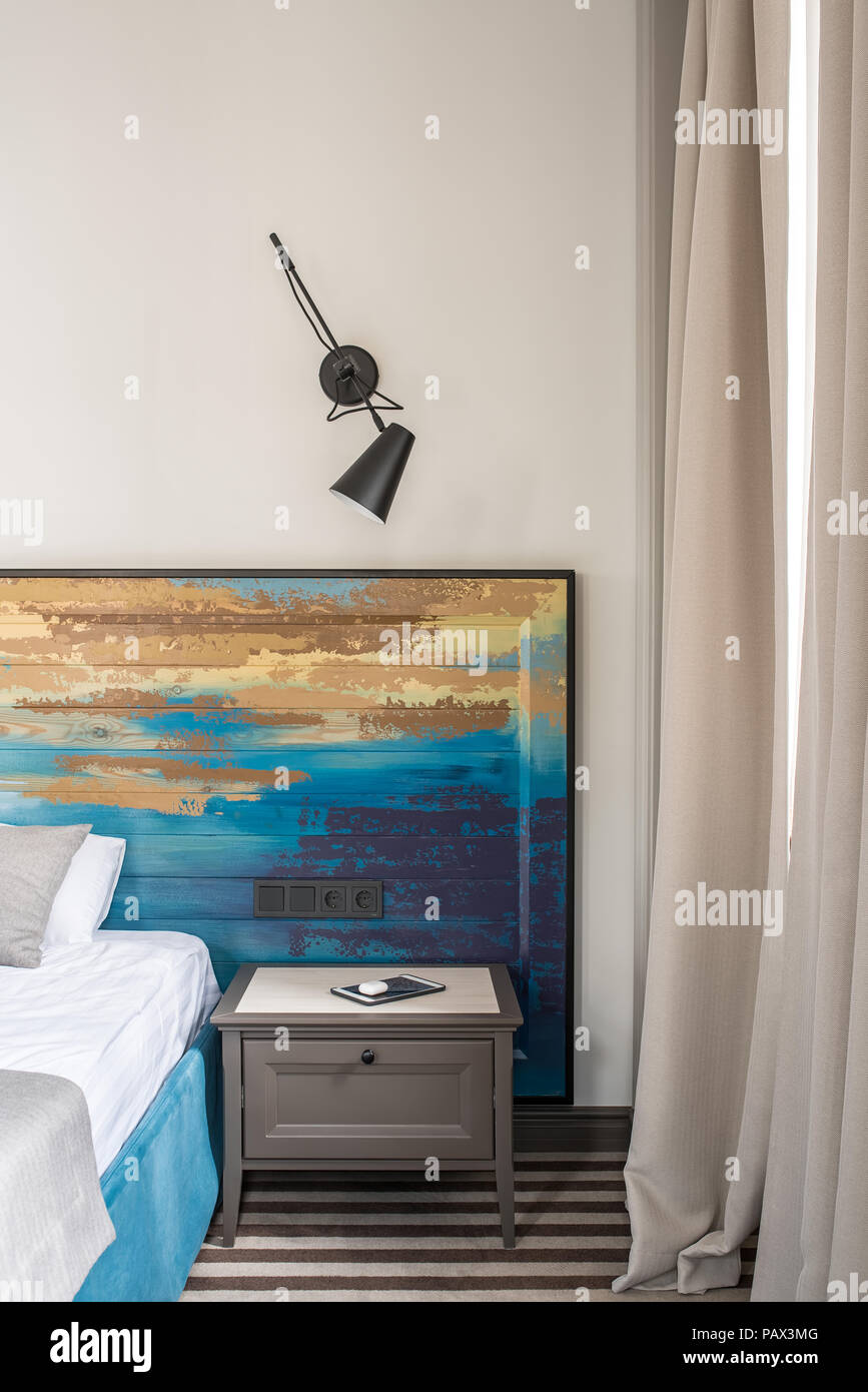 Wunderschönes Hotel Zimmer mit hellen Wänden und einem gestreiften Stock. Es ist ein blauer Bett mit einem bunten hölzernen bedhead, Nachttisch mit einer Tablette, schwarze Lampe o Stockfoto